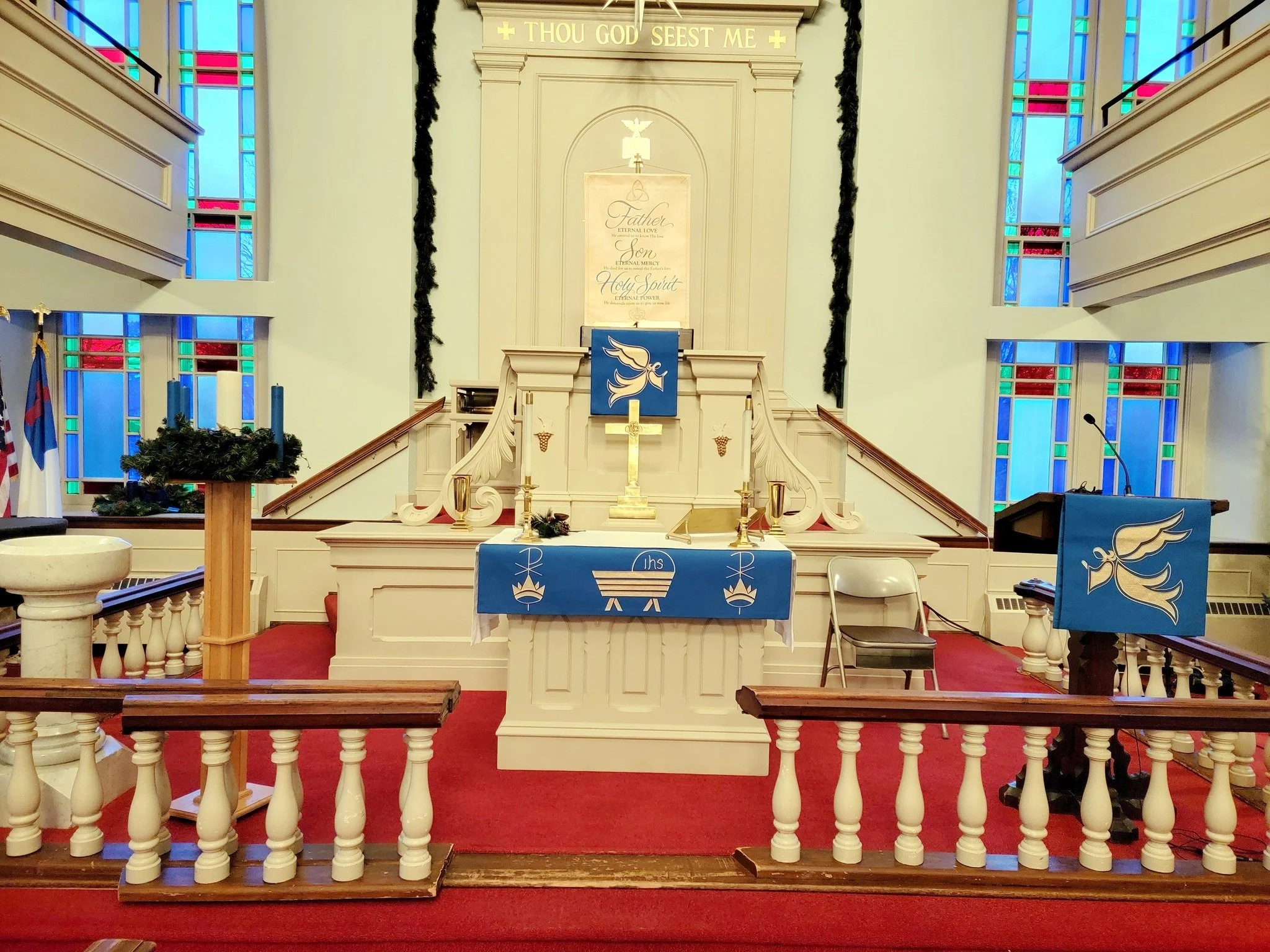 Interior of a church altar decorated for Christmas with candles, greenery, and religious symbols, stained glass windows in the background, and a blue altar cloth with a dove. A banner at the top reads 'Thou God Seest Me'. Small flags and a microphone
