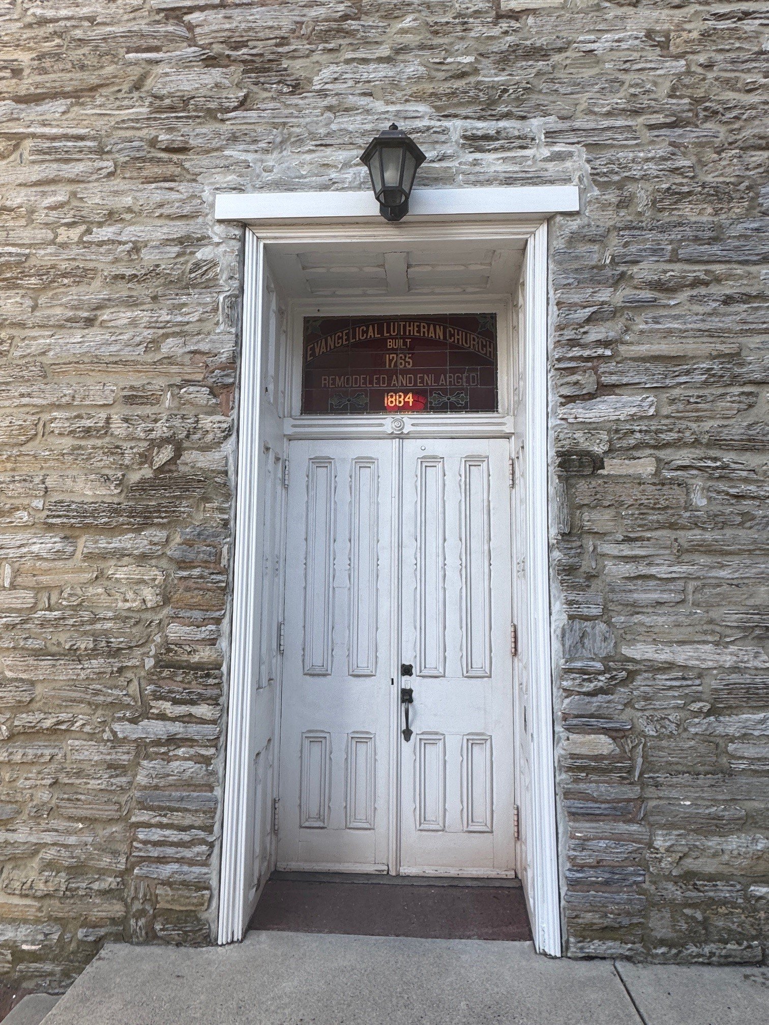 Close-up of a stone building's entrance with a white wooden door, a black lantern above, and a stained-glass window with the text 'Evanglical Lutheran Church,' 'Built 1765,' 'Remodeled and Enlarged,' and the year 1834.