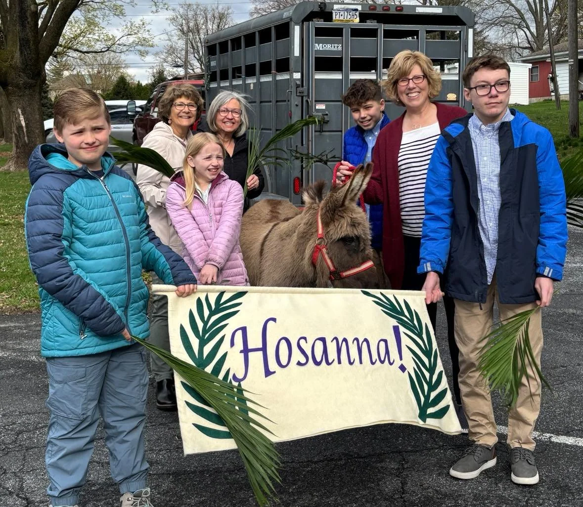 Group of smiling people, including children and adults, positioning around a small donkey holding a banner that reads 'Hosanna!' with green palm leaves. They are outdoors, standing in front of a horse trailer with a rural background.
