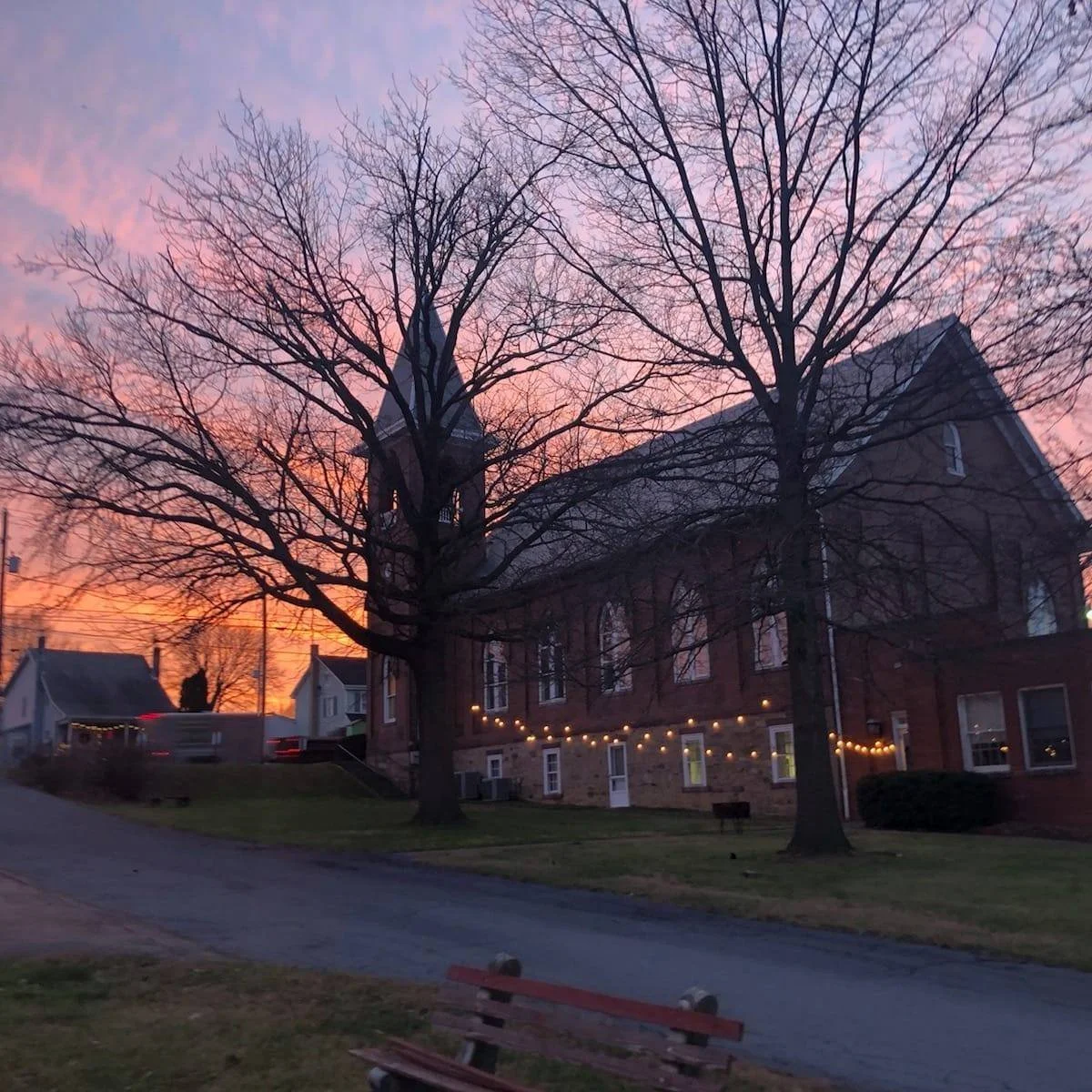 A sunset sky with pink and purple hues. Silhouettes of leafless trees in front of a large brick church building decorated with string lights, and residential houses in the background.