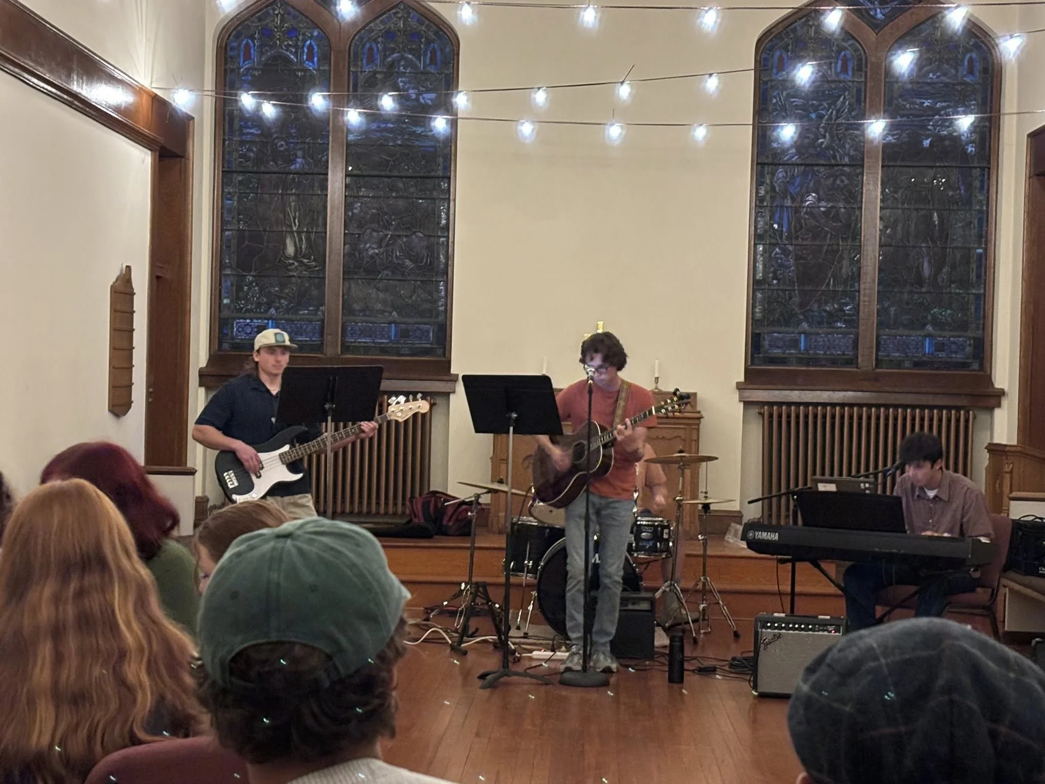 A live music performance in a church or hall with three musicians on stage. The band includes a bassist, a guitarist singing into a microphone, and a keyboardist. The stage has stained glass windows behind them, string lights hanging overhead, and an