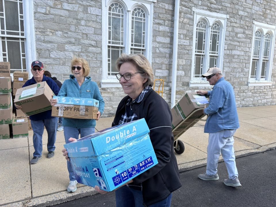 People loading boxes, including paper and supplies, outside a stone building with arched windows.