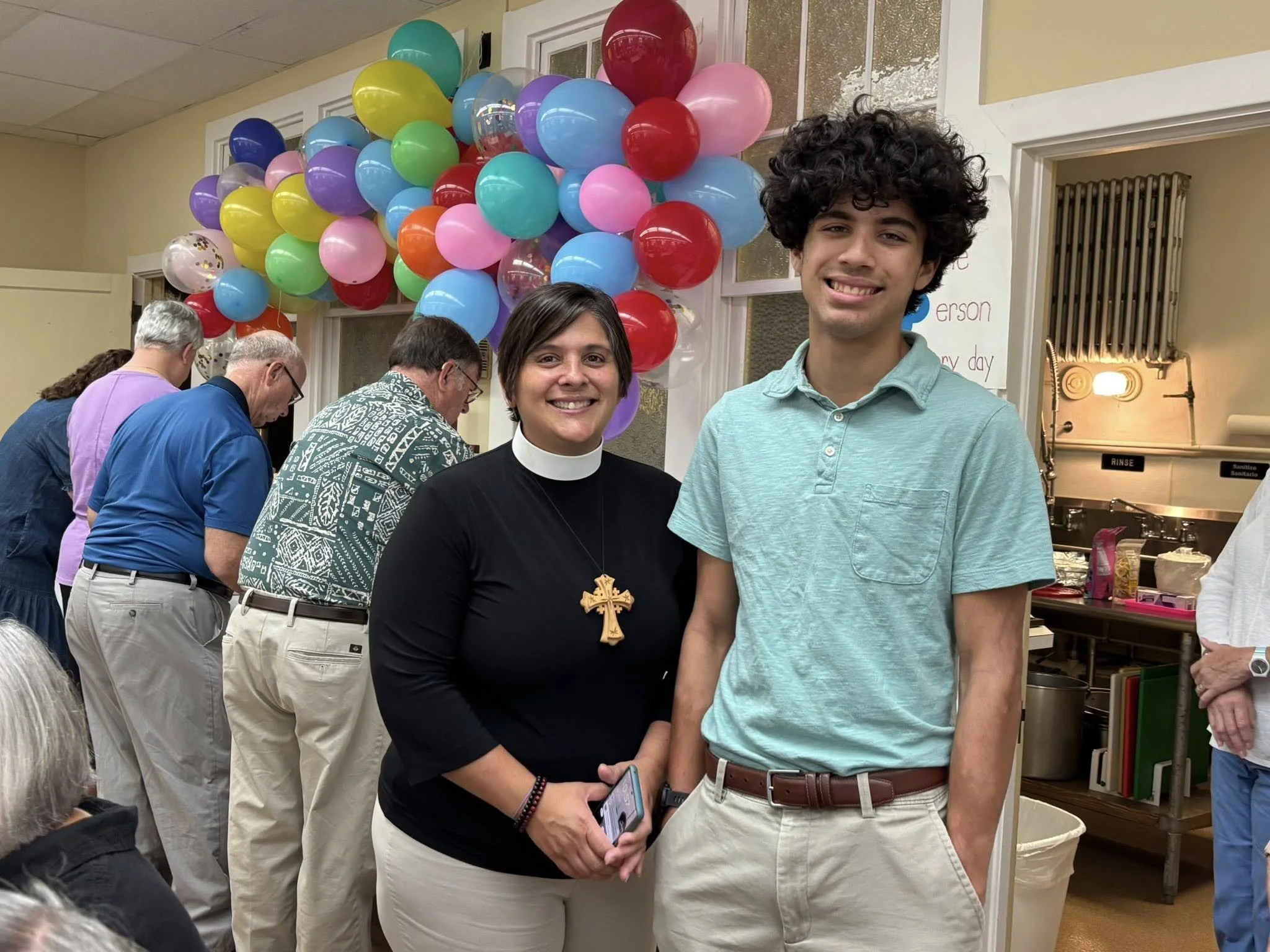 Two smiling individuals stand in front of colorful balloons at a celebration. The woman wears a black top with a clerical collar and a large cross necklace, and the young man is dressed in a light green polo shirt and beige pants. Several people are 