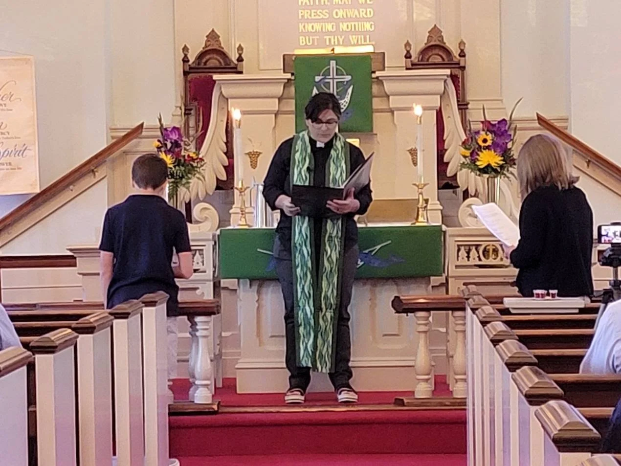 A church interior with three people, including a pastor, standing at the altar during a service. The altar has lit candles and floral arrangements. A large green cloth with a Christian symbol hangs behind them, and a message is displayed above.