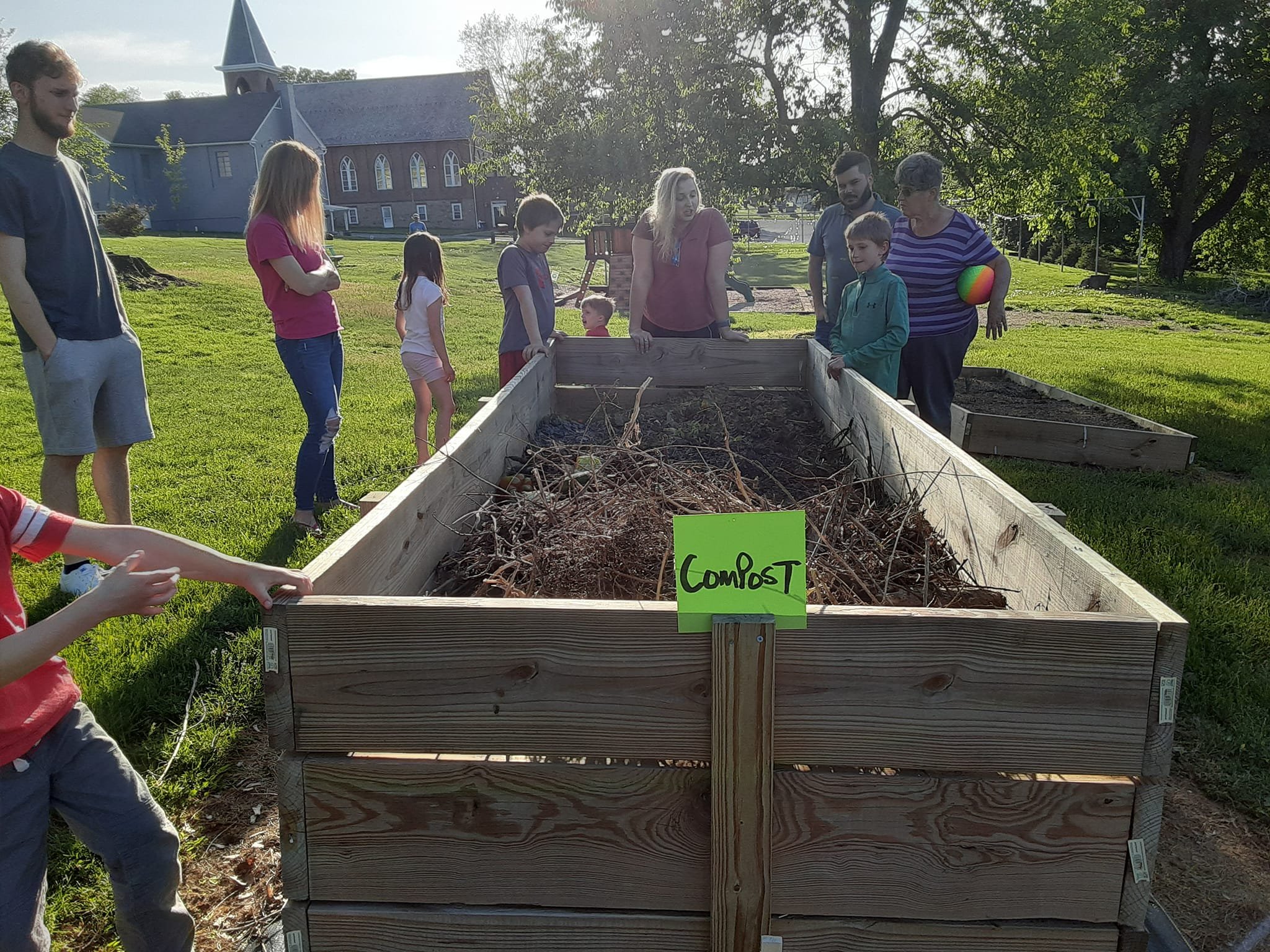 Children and adults gathered around a large wooden compost bin outdoors on a sunny day, with a church and trees in the background.