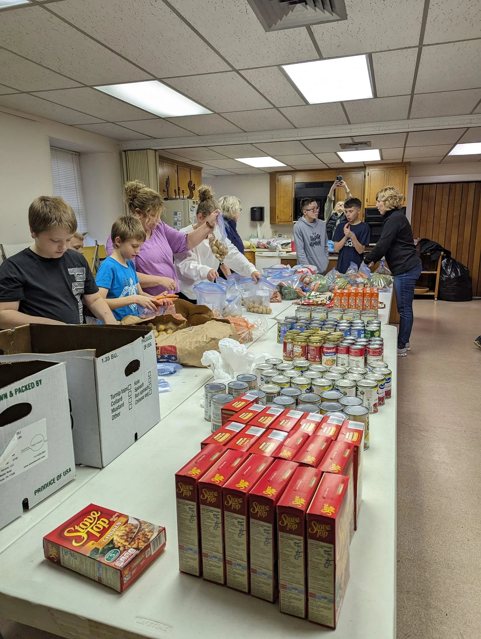 People, including children, preparing and packing food items on a table for a community service or food drive event, with canned goods, boxed food, and supplies visible.