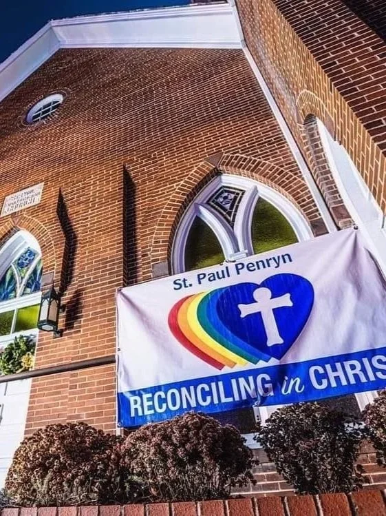 A brick church with a stained glass window, a hanging lantern, and a banner that reads "St. Paul Penryn RECONCILING in CHRIST" with a rainbow-colored heart and a white cross inside it.