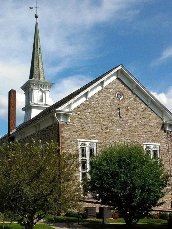 The image features a brick church with a steeple and a weather vane, surrounded by green trees and a partly cloudy sky.