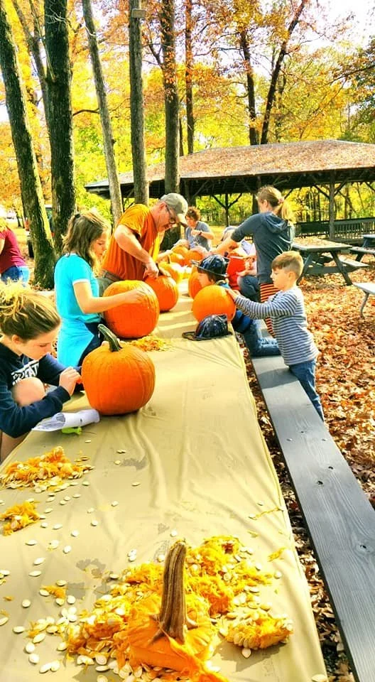 Children and adults carving pumpkins on a long outdoor table during fall, with trees showing autumn leaves in the background.