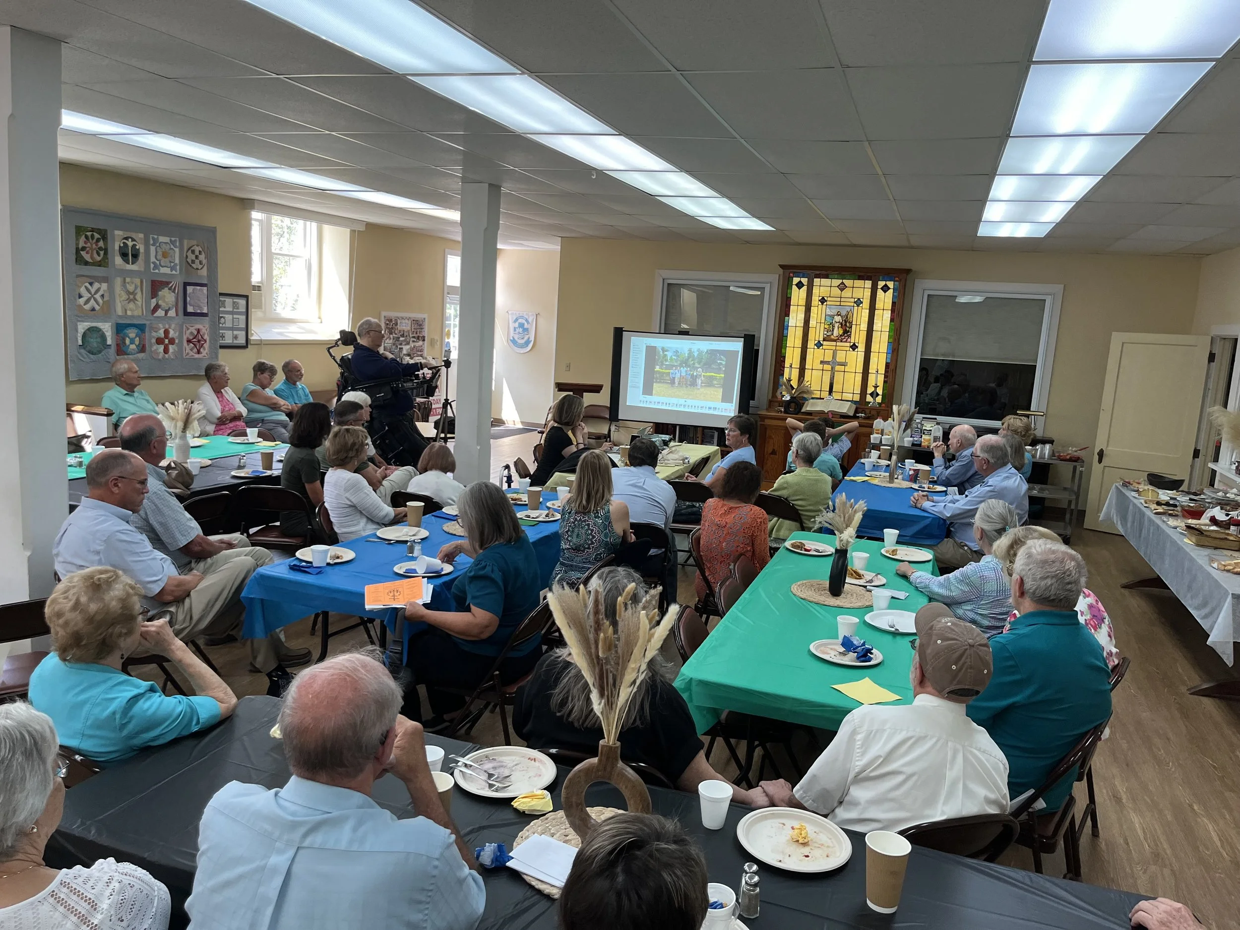 A group of people gathered in a community room watching a presentation on a screen, with tables set for a meal and a stained glass window in the background.
