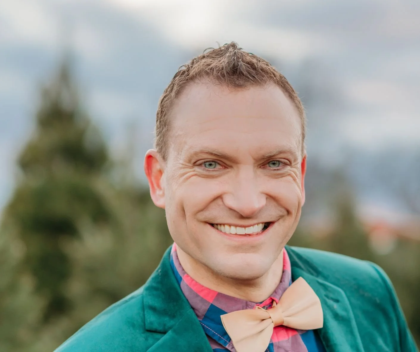 A man with short, dark blond hair and glasses, wearing a bow tie, with blurry trees in the background.