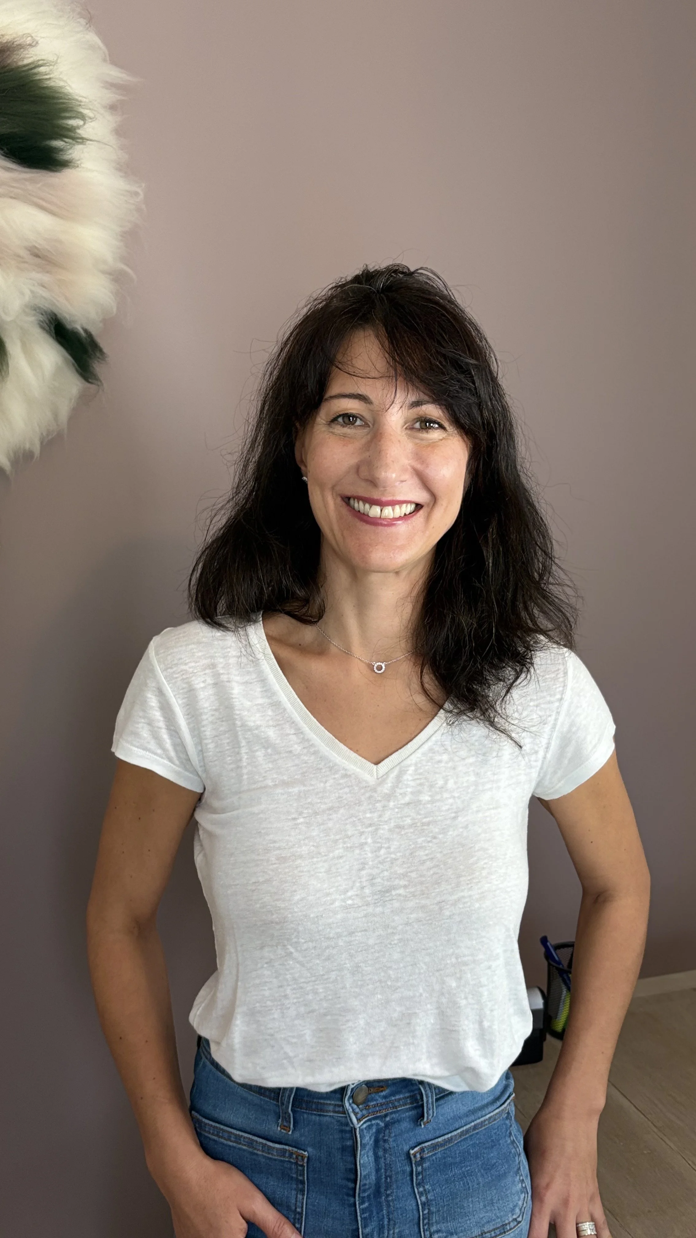 Sandra, une femme souriante avec des cheveux bruns ondulés porte un t-shirt blanc et un jean, debout contre un mur beige.