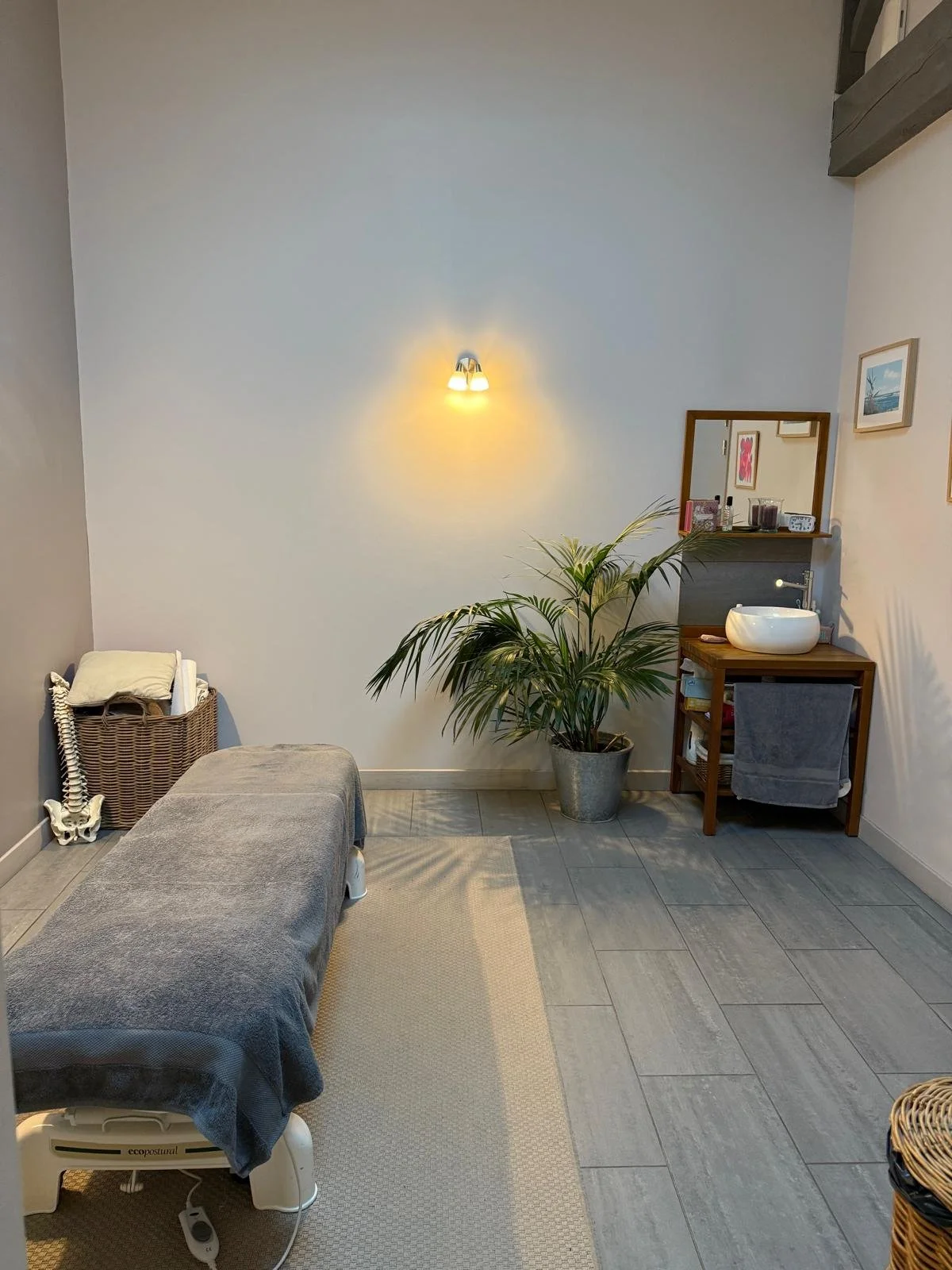 Salle de massage avec un lit de massage recouvert d'un drap gris, une plante d'intérieur, une petite table avec un lavabo en céramique, un miroir, des cadres photo, un panier à linge, un petit meuble en bois avec des serviettes et des objets décoratifs.