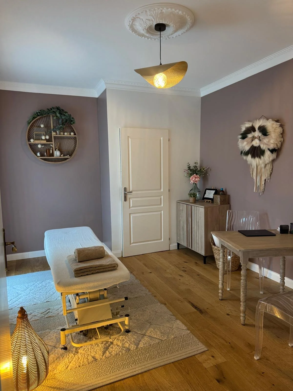 Une salle de consultation ou de massage avec un lit, une lumière jaune suspendue, un mur violet avec une décoration en forme de nuage en peluche, une armoire en bois clair avec des fleurs, une table en bois clair avec une planche, deux chaises transparentes, un tapis beige, et une lampe de sol