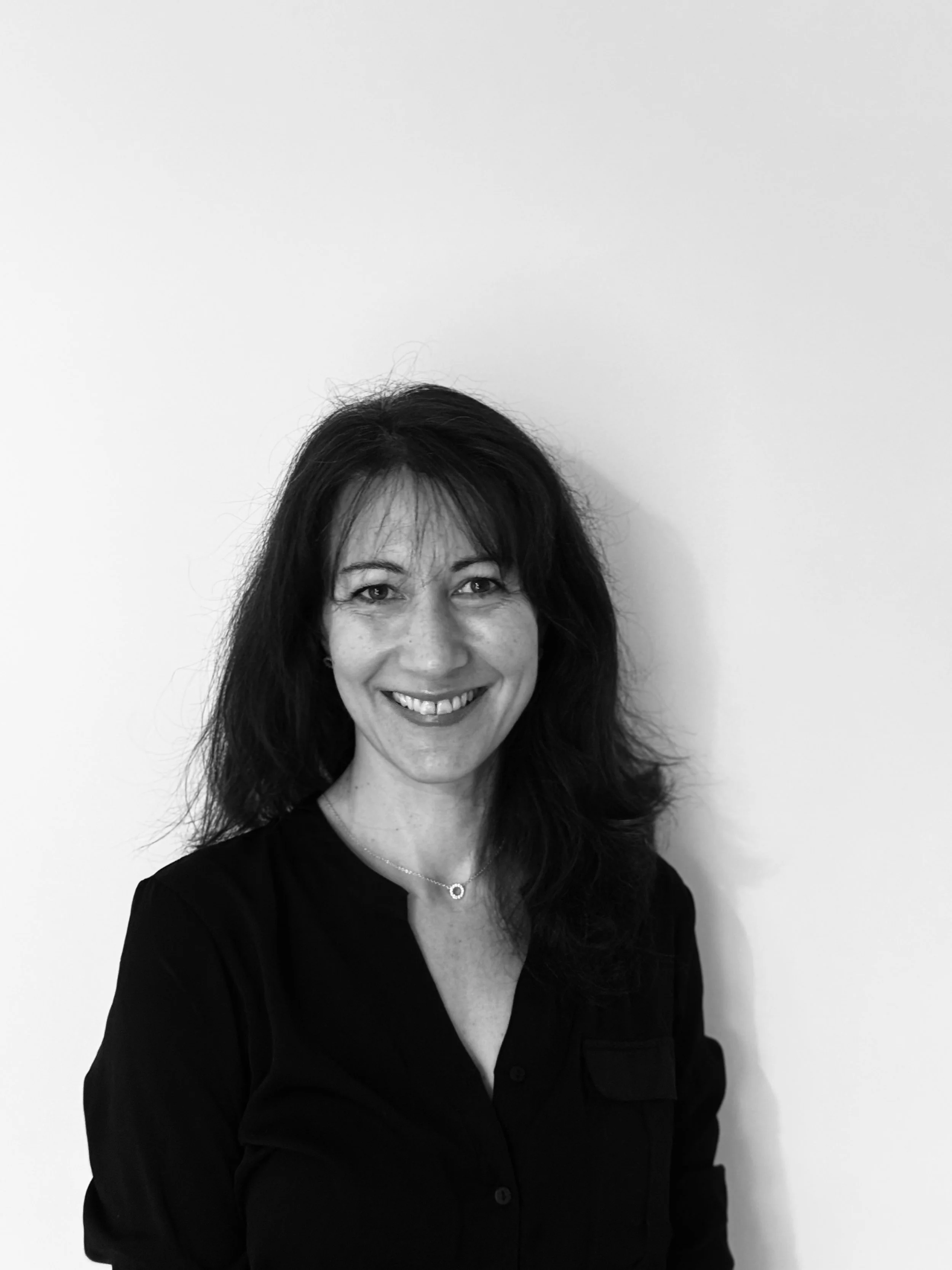 Photo en noir et blanc d'une femme souriante avec des cheveux longs, portant un chemisier noir, se tenant contre un mur blanc