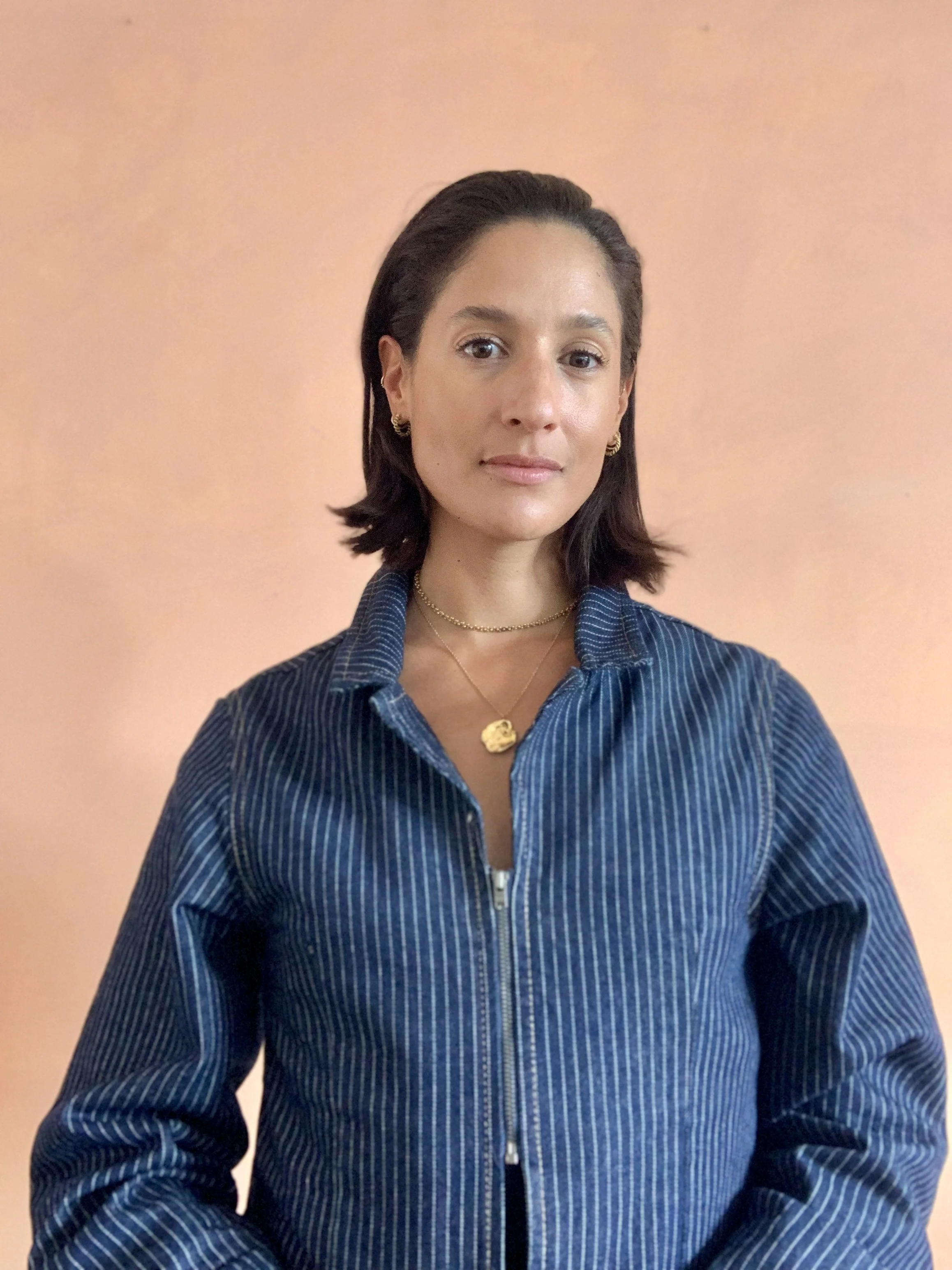 A woman with shoulder-length dark hair and jewelry, wearing a blue pinstriped zip-up jacket, standing against a plain peach-colored wall.