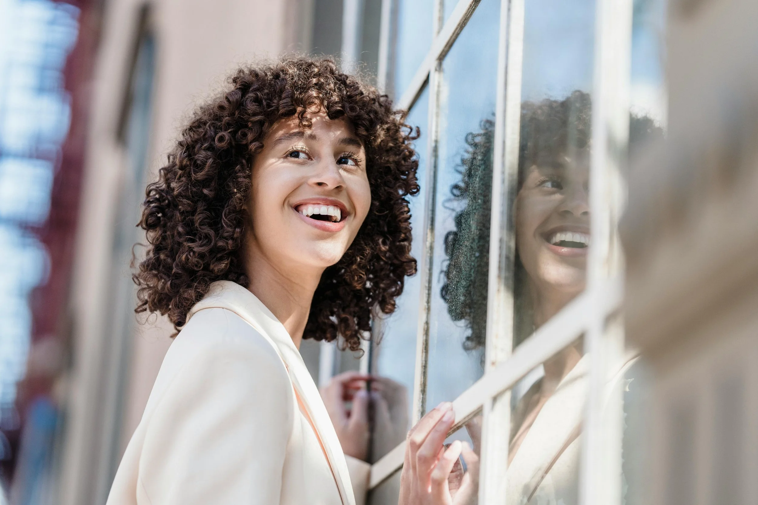 A young woman with curly hair smiling and looking away while standing next to a window with a reflection of her face in the glass.