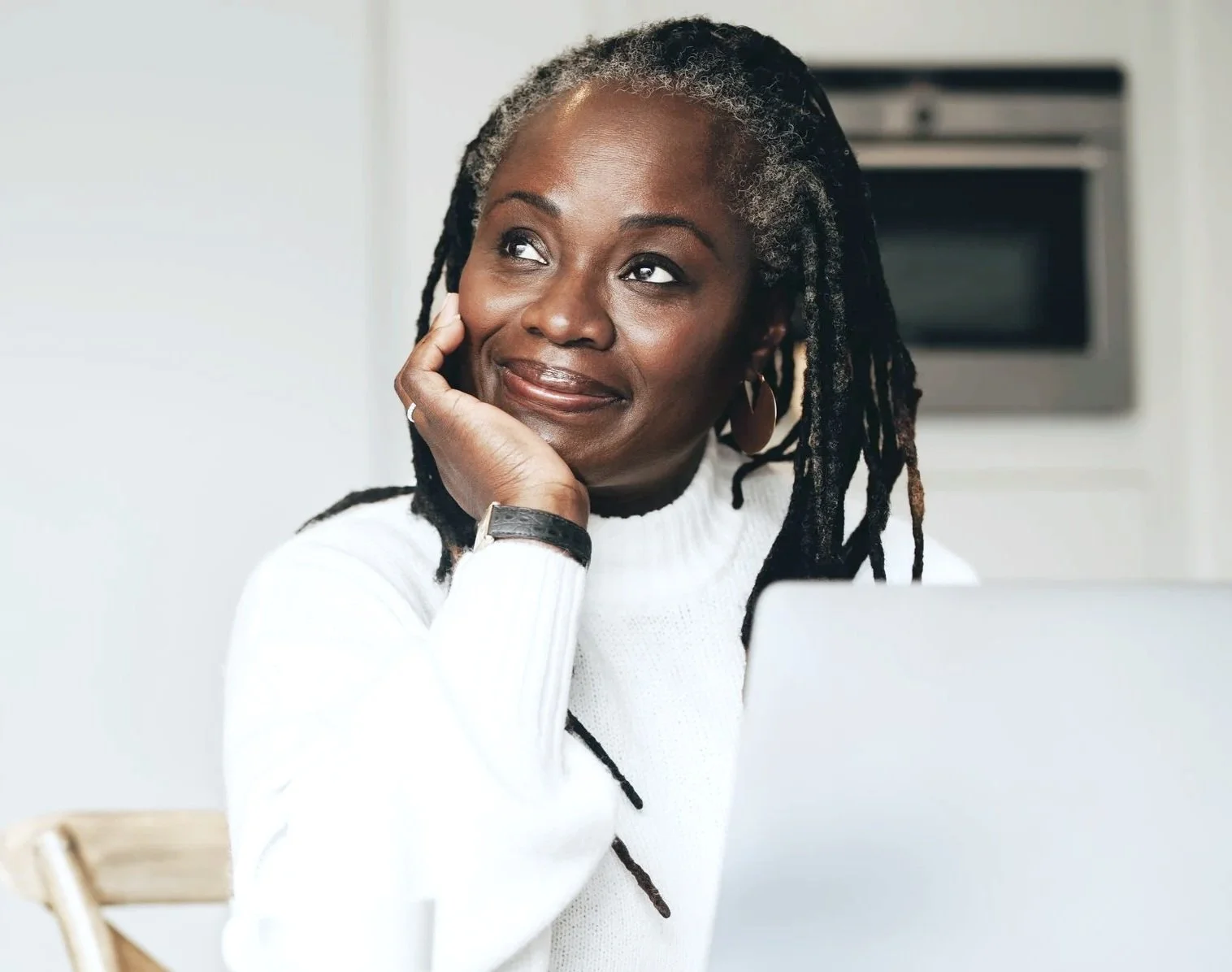A woman with gray dreadlocks and dark skin, smiling thoughtfully while resting her chin on her hand, sitting in front of a laptop.