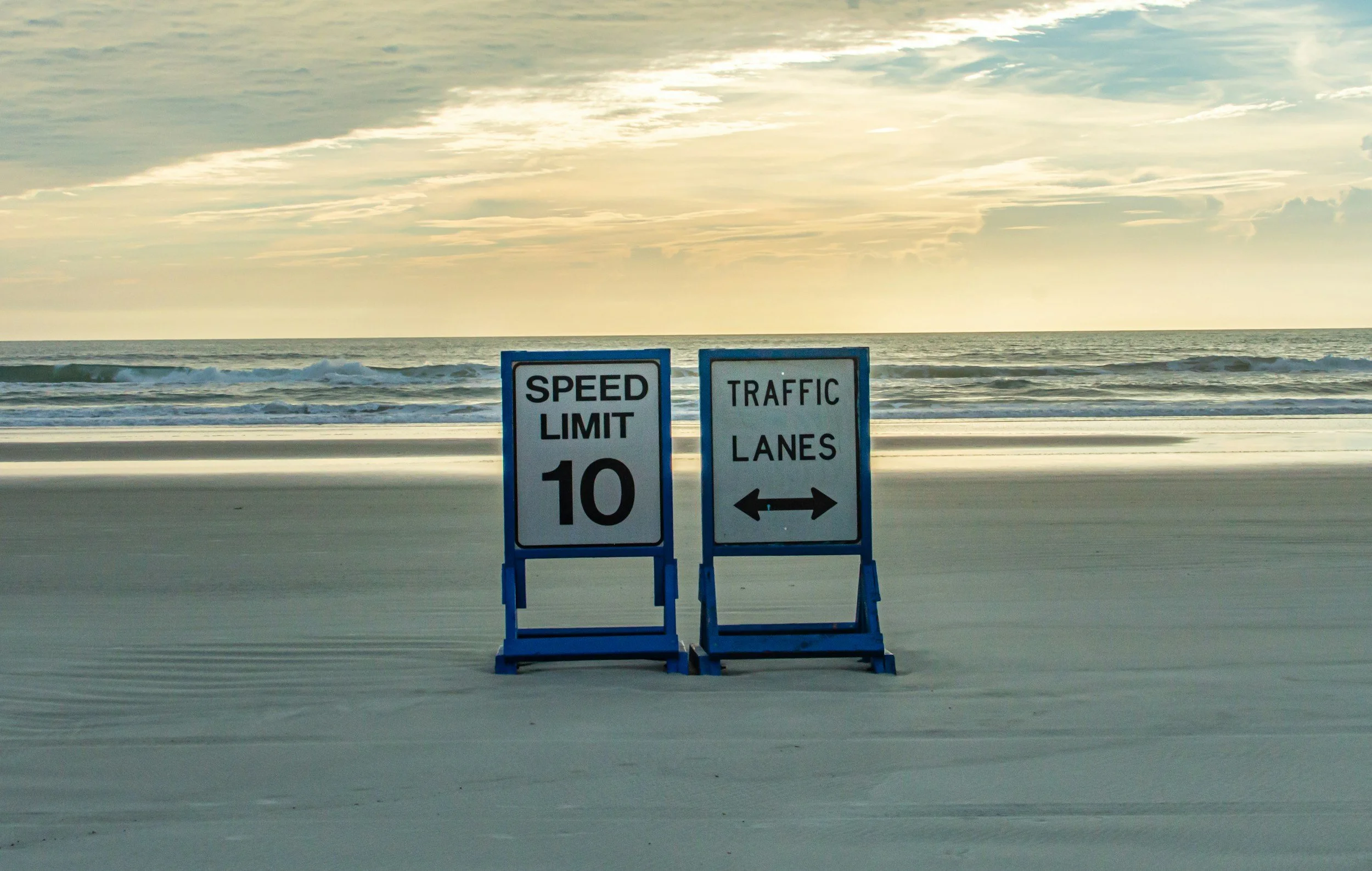 Two blue traffic signs on the beach, one indicating a speed limit of 10 and the other directing traffic lanes both ways, with the ocean and sky in the background.
