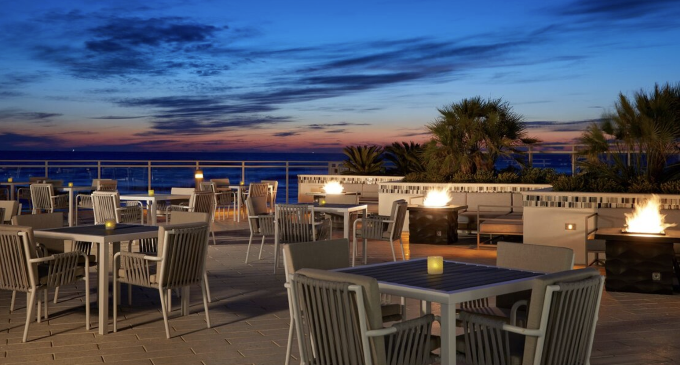 Outdoor rooftop dining area at sunset with fire pits, tables, chairs, and decorative plants.