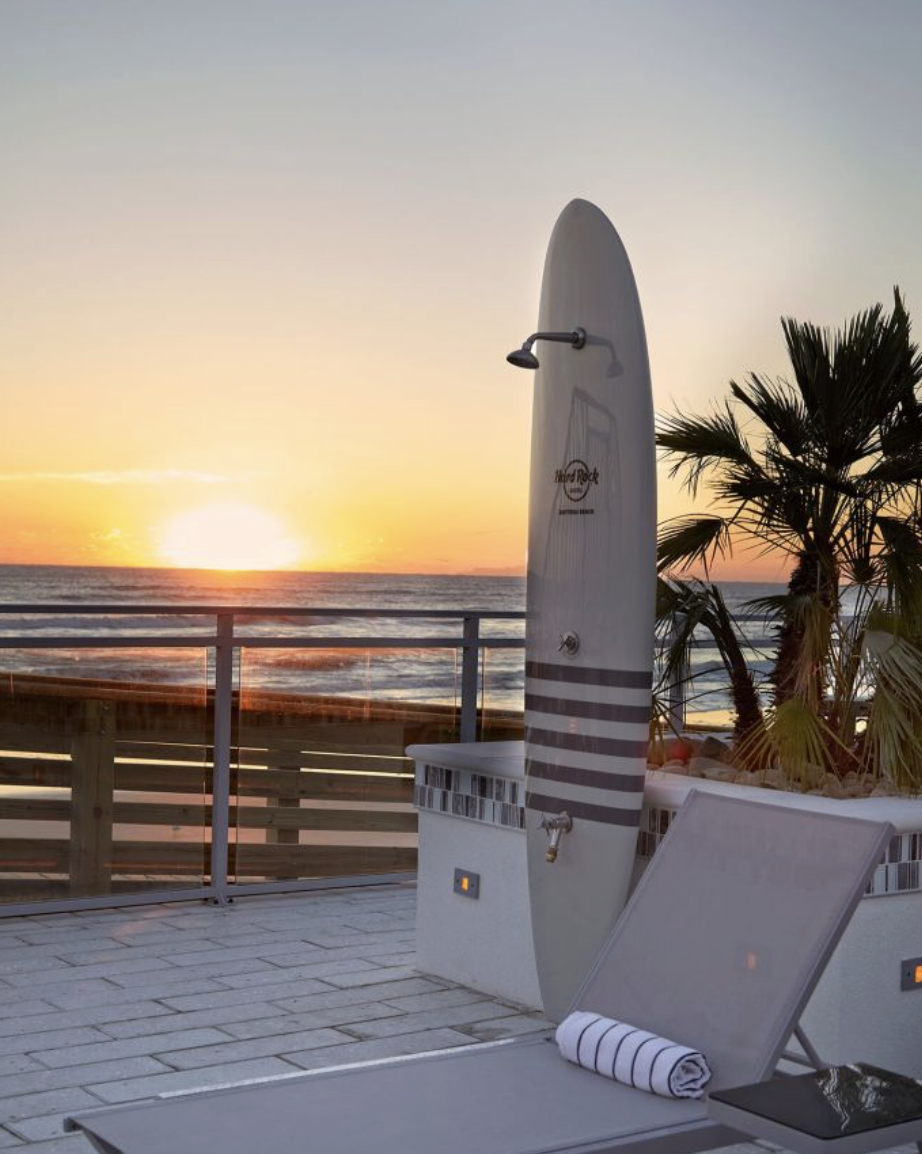 Sunset over the ocean with an outdoor shower and a lounge chair on a patio, palm trees in the background.