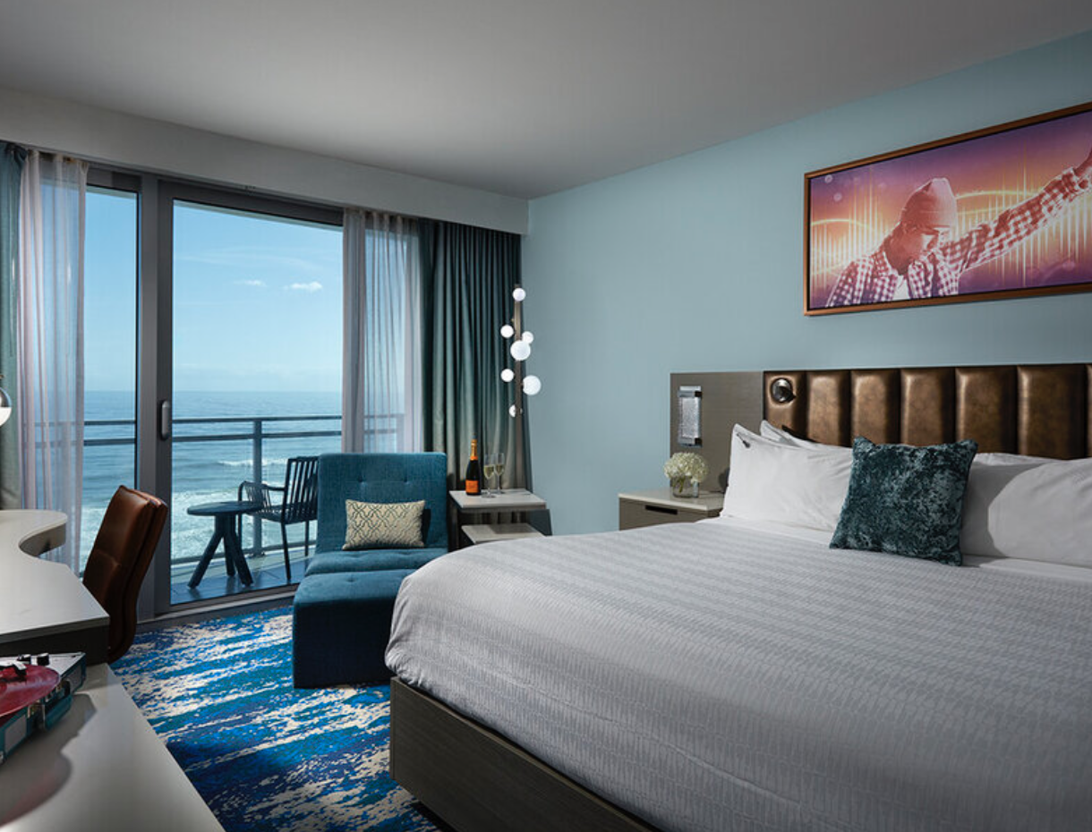 A hotel room with a ocean view, featuring a bed with white sheets, blue decorative pillows, a brown headboard, a white nightstand with flowers, a bottle of champagne, and a glass. There is a blue armchair with a pillow, a small table, and a balcony with outdoor seating overlooking the ocean. A colorful abstract digital art piece is on the wall.