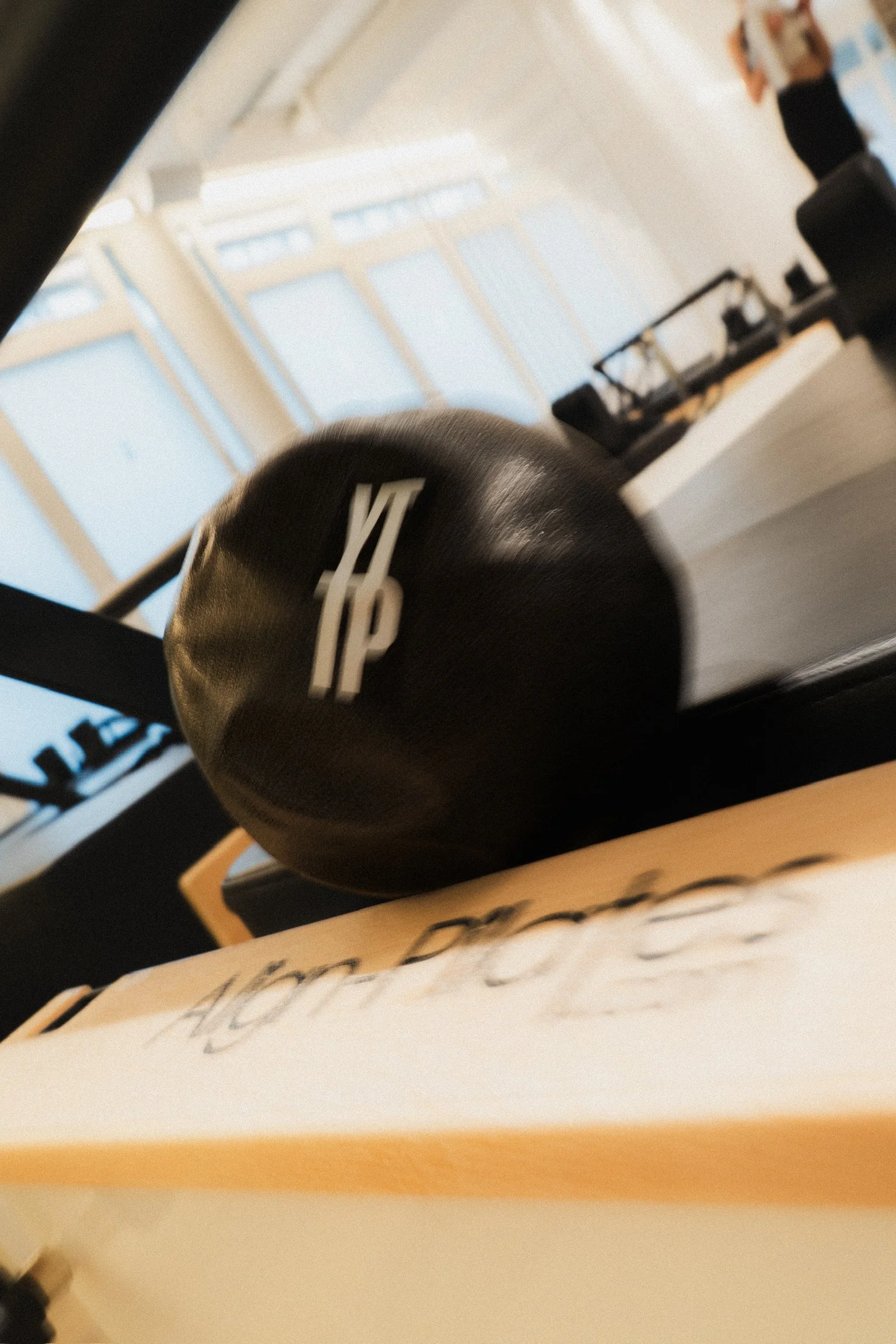 Ein blacker Medizinball mit weißer Aufschrift befindet sich auf einem Tisch in einem Raum mit großen Fenstern und Hängeschildern.