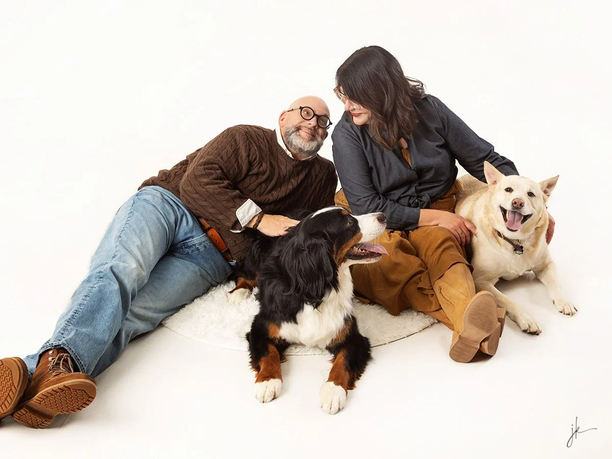 A man with glasses and a beard, and a woman with dark hair, sitting on the floor with two dogs in front of a plain white background. The man is lying on his side, the woman is sitting cross-legged, and they are smiling at each other. The dogs are happy and relaxed, one is a black and white Australian Shepherd, and the other is a cream-colored Labrador Retriever.