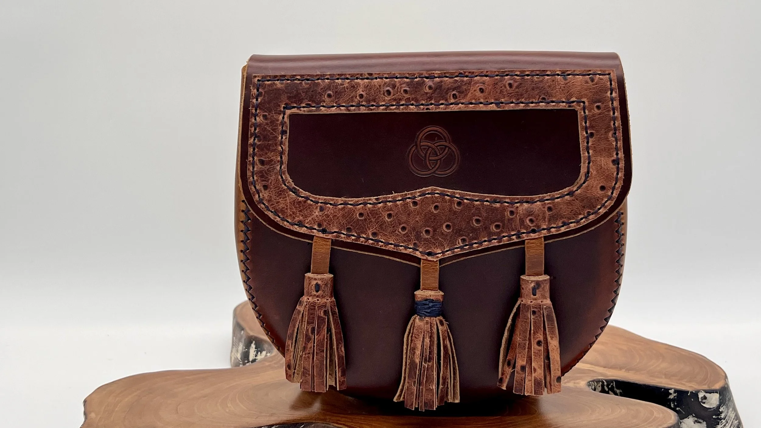 A leather handbag with a flap closure, decorated with three tassels hanging from the front, placed on a wooden surface against a plain background.