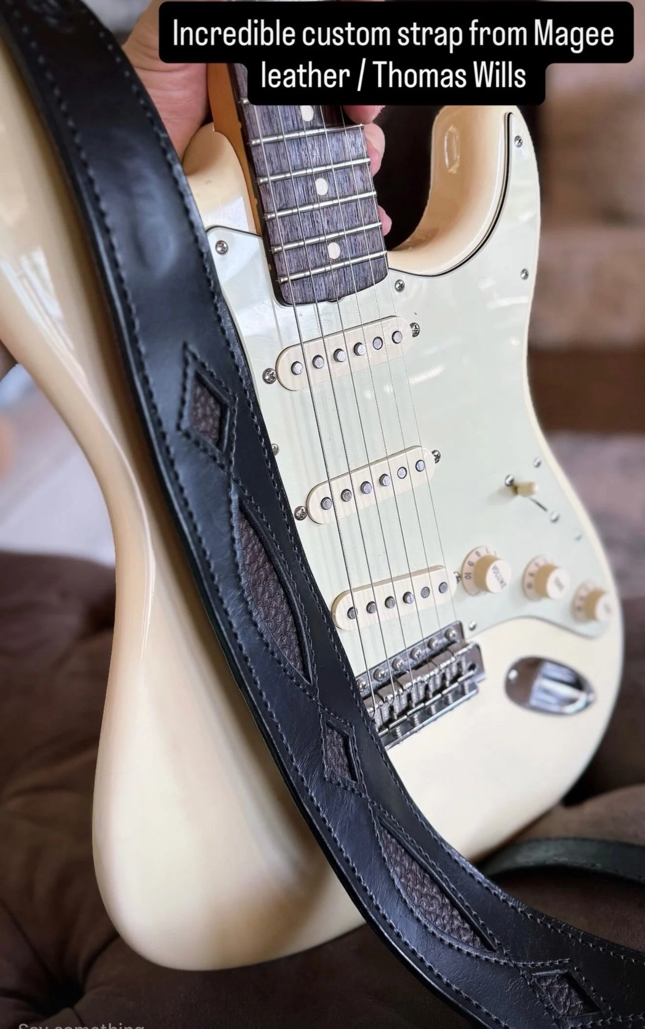 A cream-colored electric guitar with a black leather strap featuring diamond-shaped quilted leather accents. The guitar has a white pickguard, three single-coil pickups, and cream-colored knobs and a tone switch. The image shows a close-up view of th