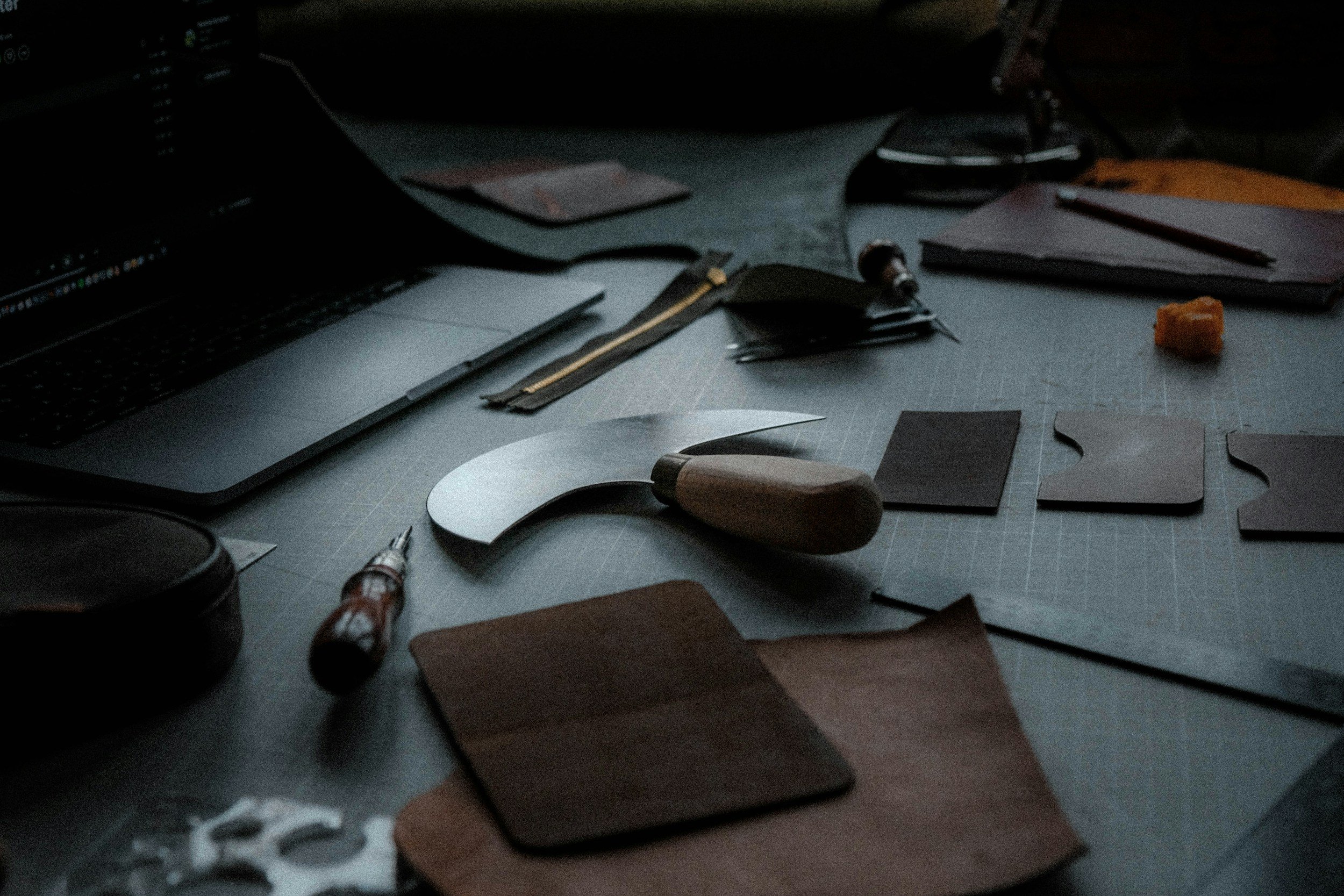 Workstation with leatherworking tools, pieces, and a laptop on a dark desk.