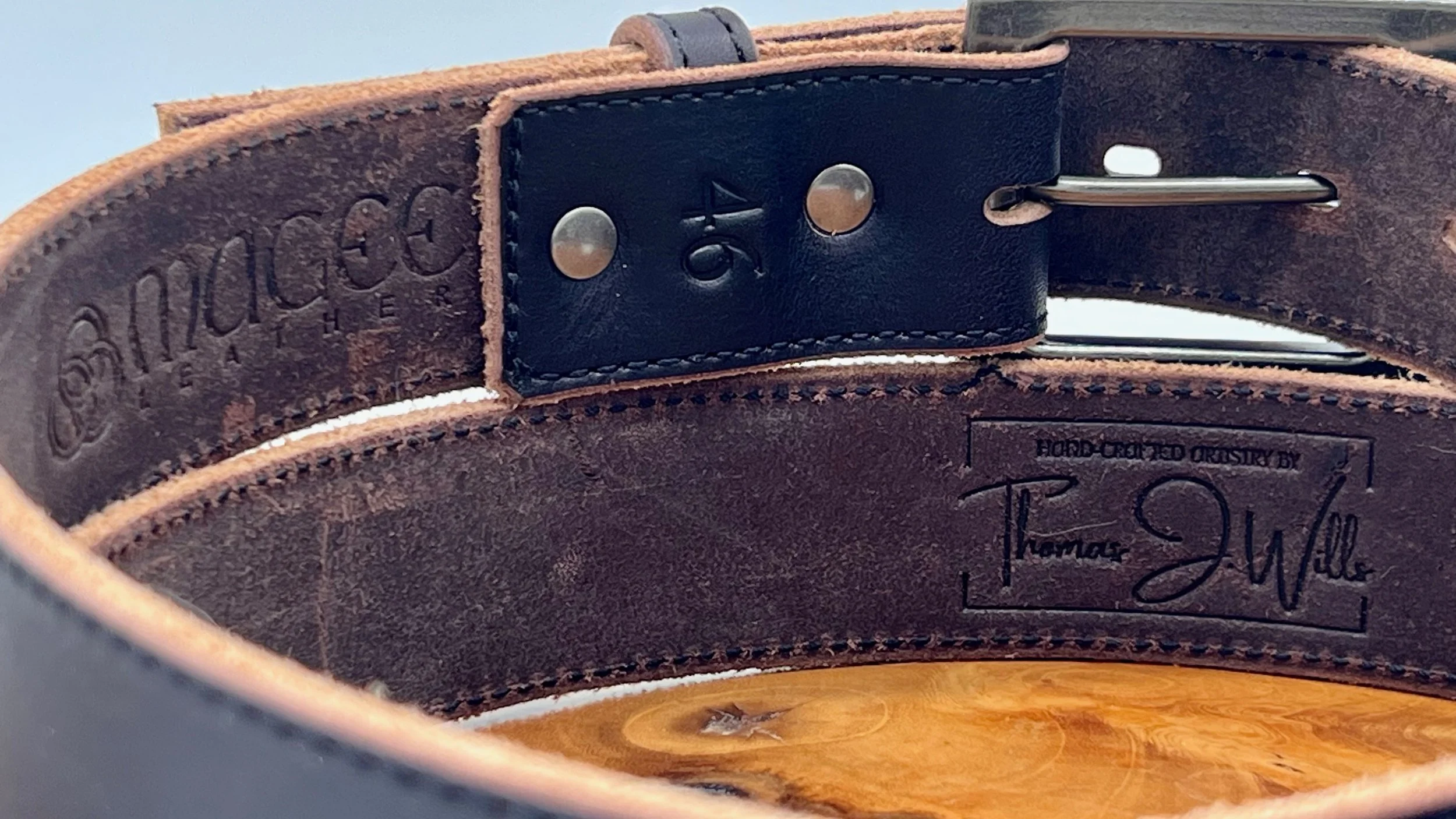 Close-up of a brown leather belt with embossed brand name and date, featuring a black leather patch and a metal belt buckle.
