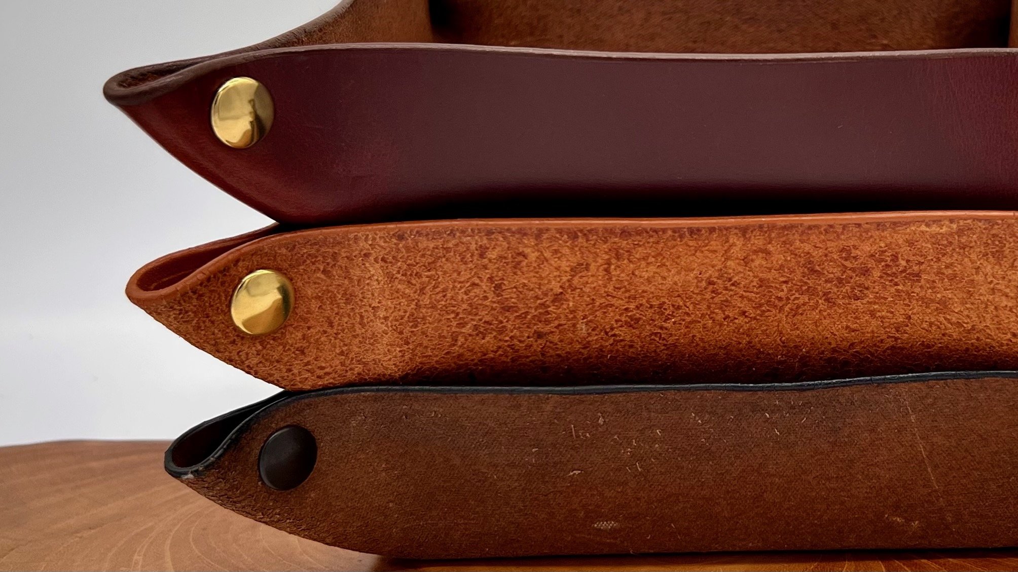 Close-up of three stacked leather folders or books with pointed ends and metal or plastic caps at the tips, in shades of brown and red, on a wooden surface.