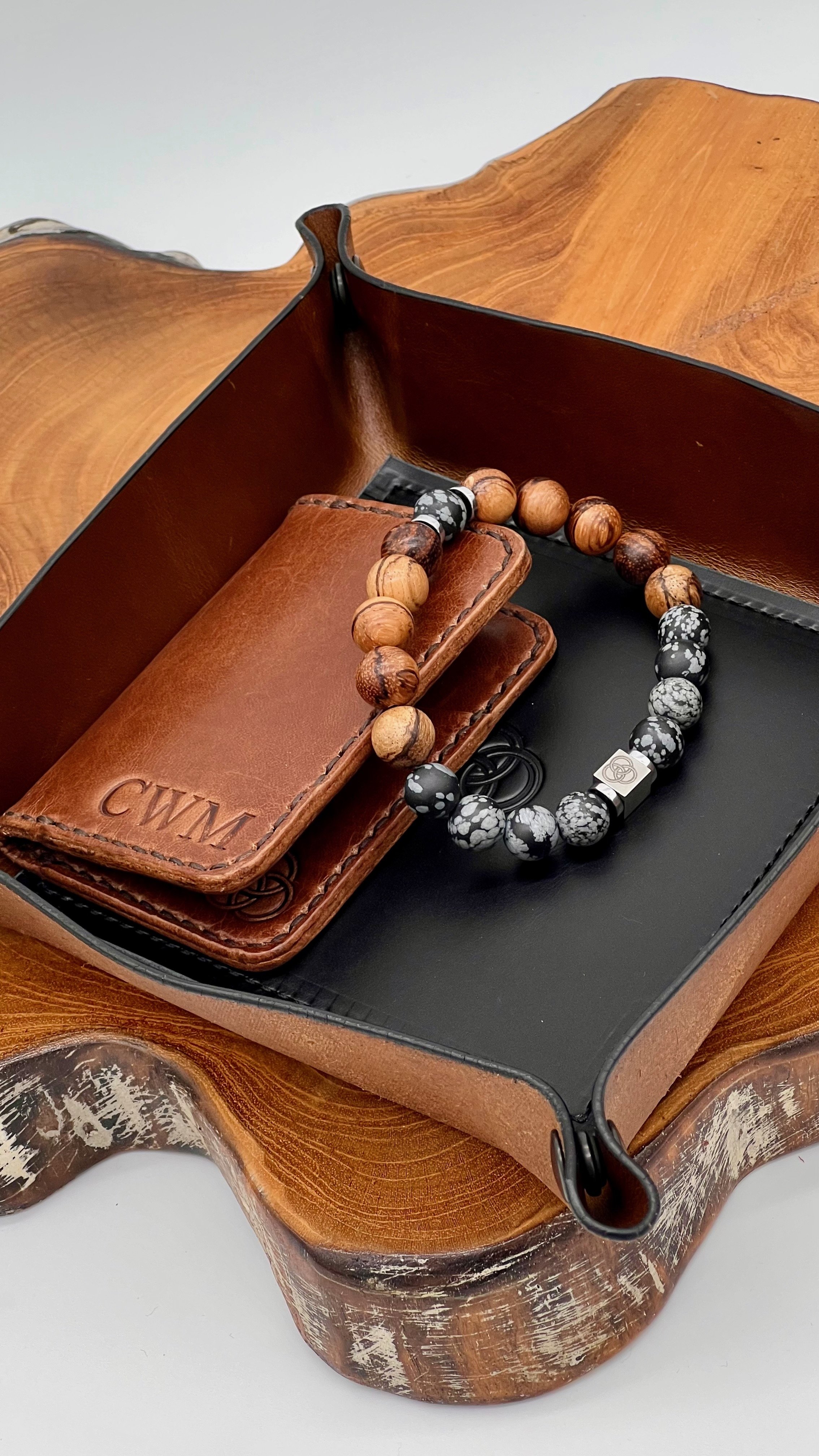 A beaded bracelet with a mix of black, white, and tan beads, and a metal square charm, placed inside an open brown leather box on a wooden surface.