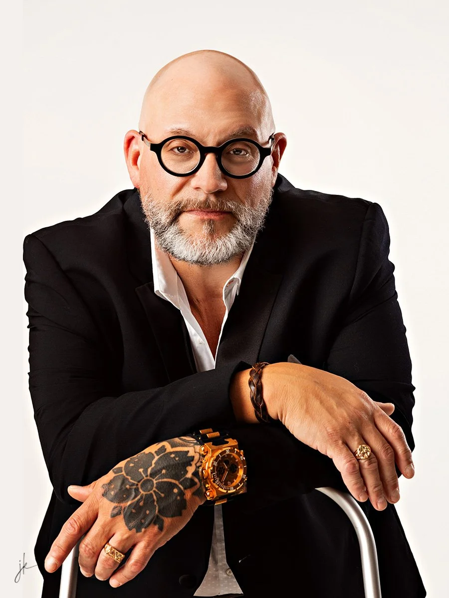 A bald man with a beard wearing black-rimmed glasses, a black suit, and a white shirt. He has a large tattoo of flowers on his left hand, an orange watch, and a ring on his right hand. He is resting his arms on a white cane and looking at the camera with a neutral expression.