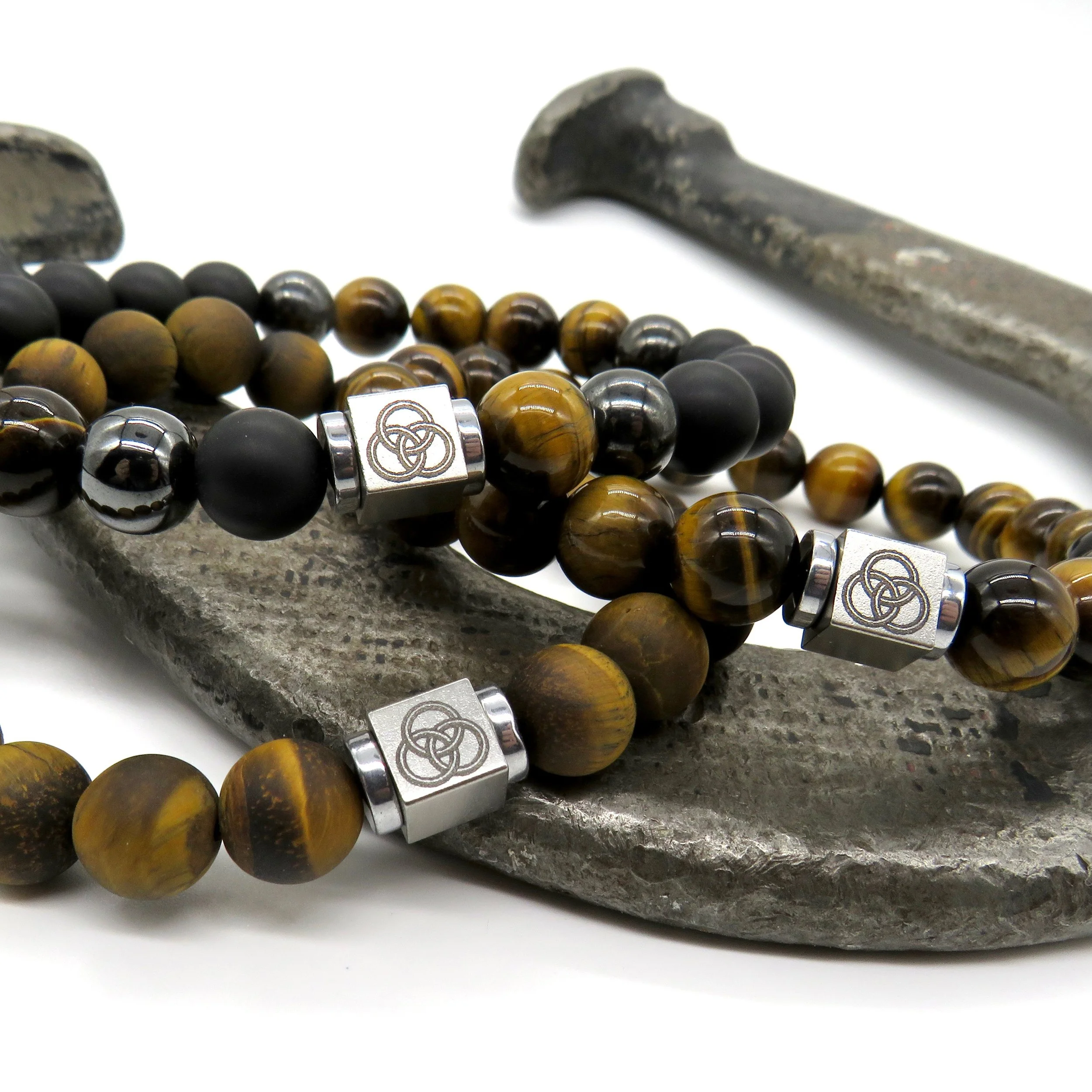 Bracelet with brown and black beads and silver accents featuring interlocked rings, resting on a rough metal surface with a metal nail nearby.