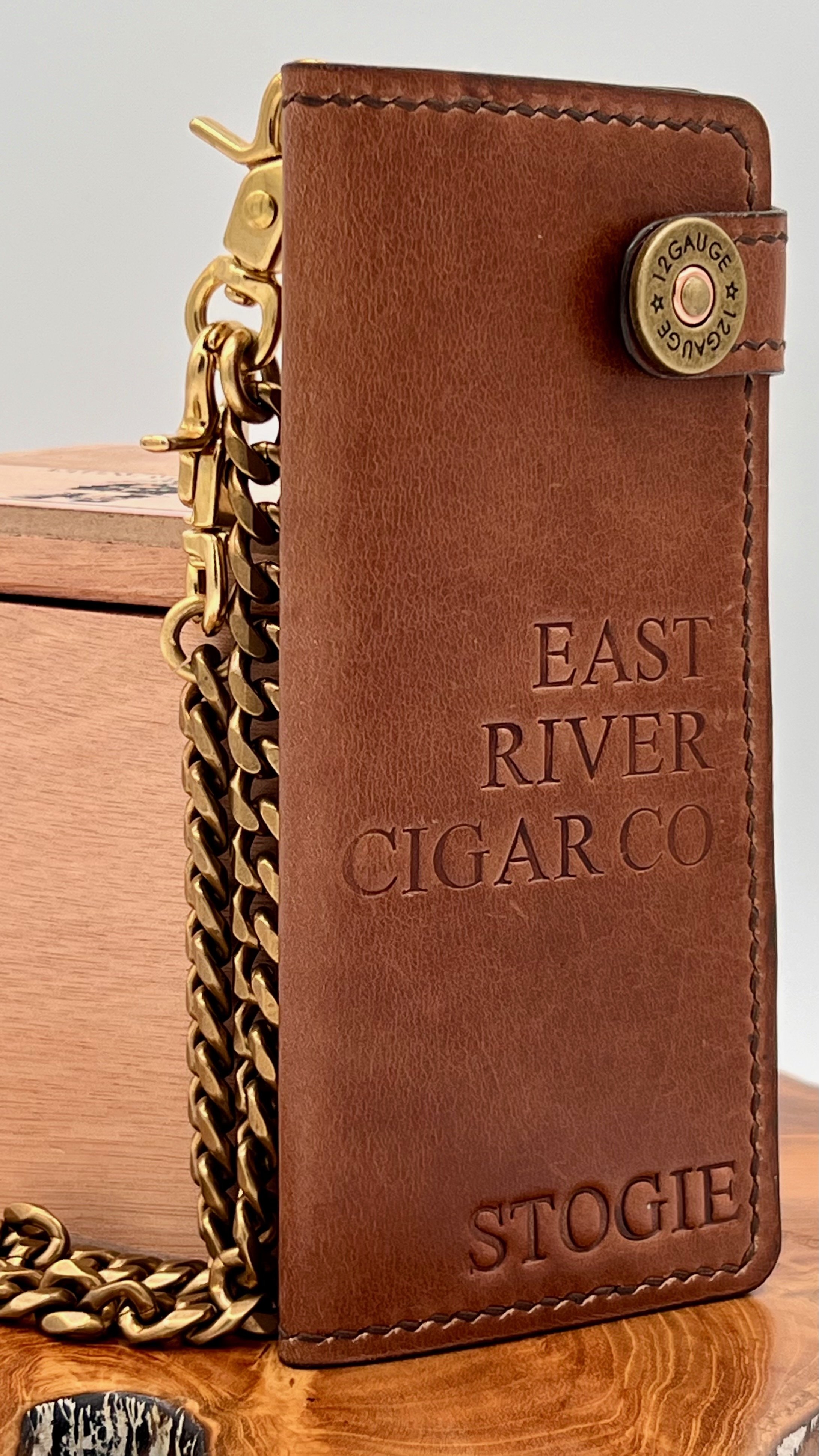 Leather cigar case with engraved text 'East River Cigar Co. Stogie', attached to a gold chain, with a bullet-shaped clasp reading '12 Gauge'.