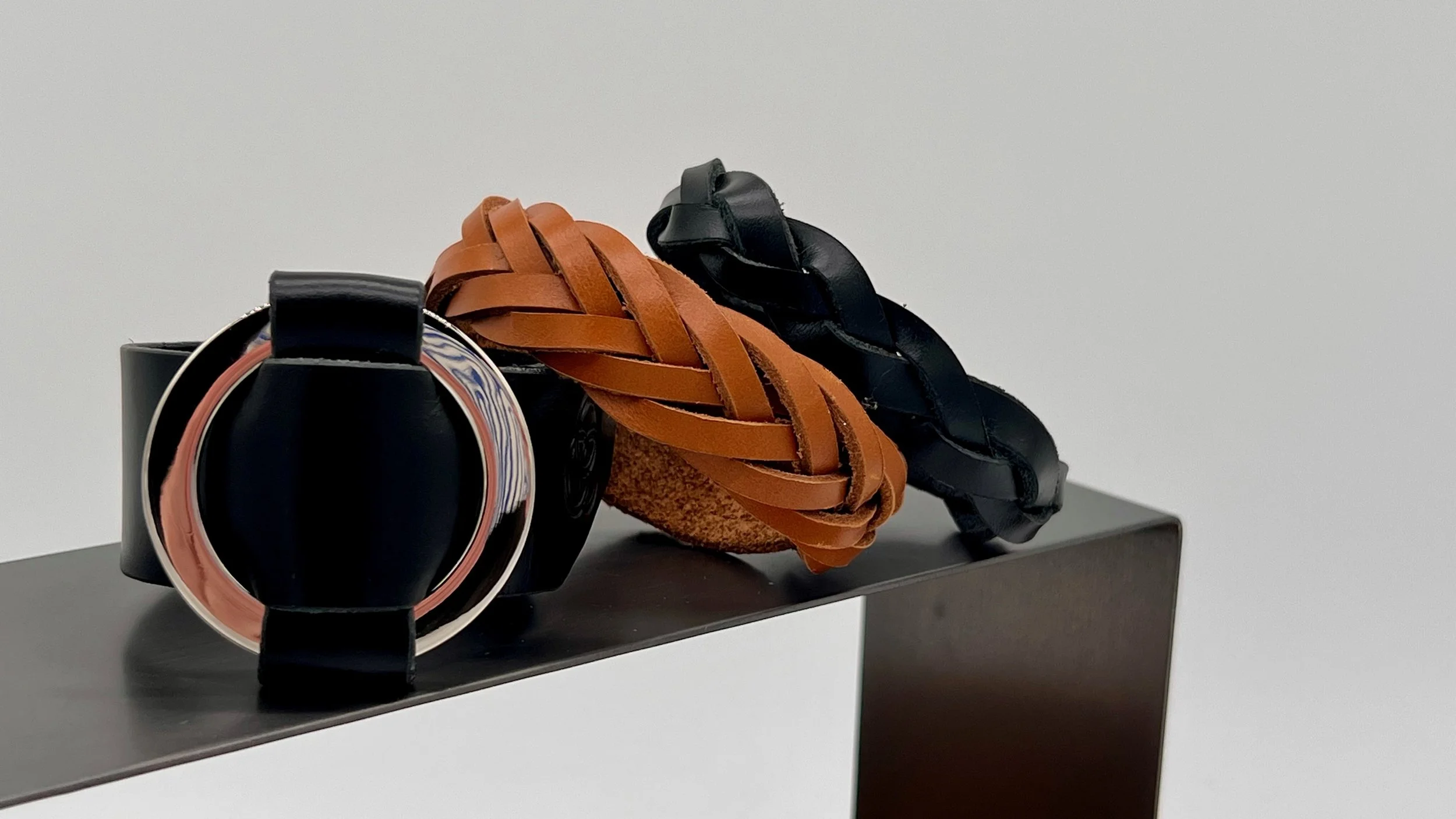 A black watch, a brown braided leather headband, and a black braided leather headband, all displayed on a black stand against a white background.
