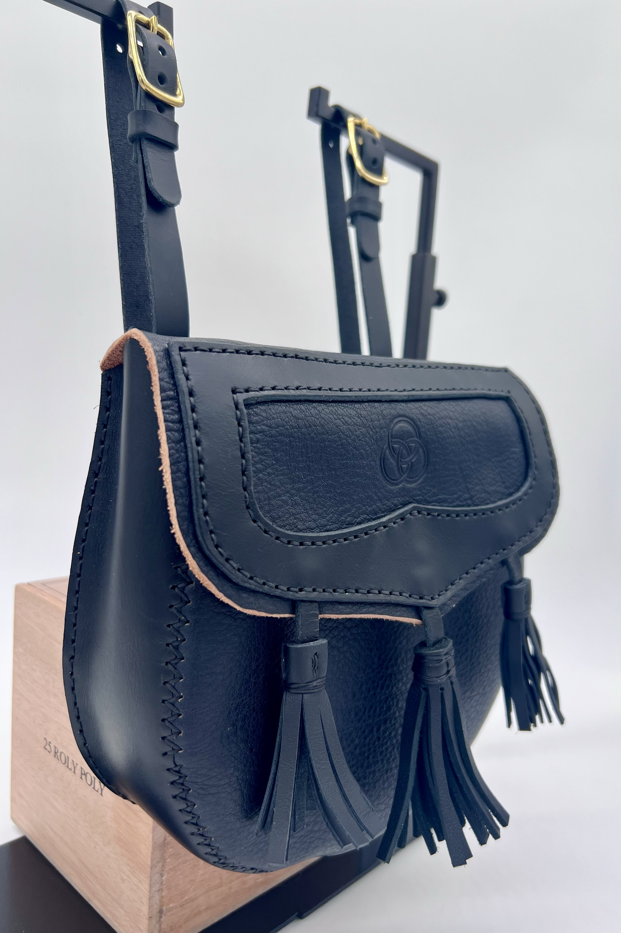 Close-up of a black leather handbag with tassel decorations hanging from the front, placed on a wooden box with the words '25 BOLLY POLY'. In the background, there are two adjustable black metal stands or fixtures with gold buckles, against a plain l