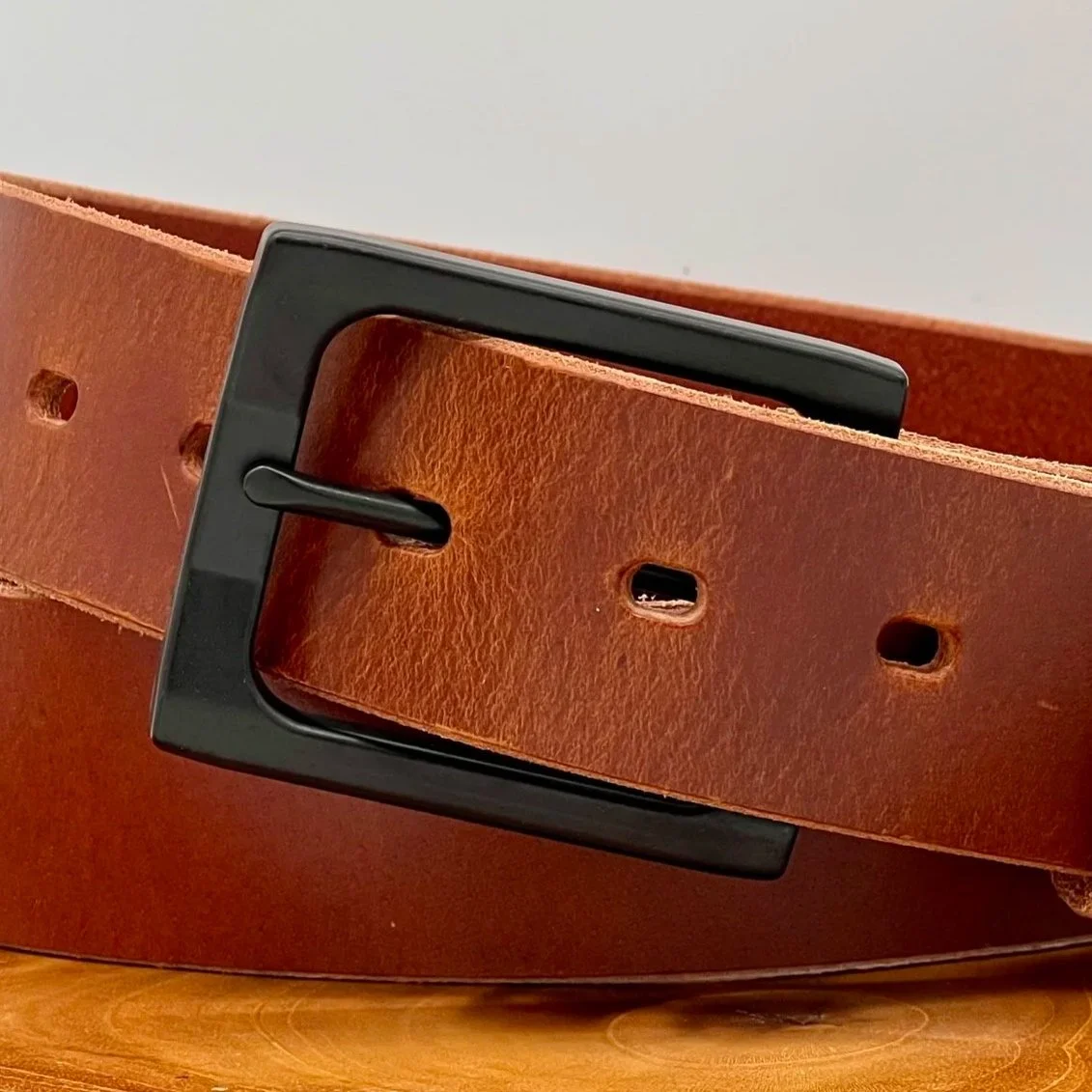 Close-up of a brown leather belt with a black rectangular buckle resting on a wooden surface.