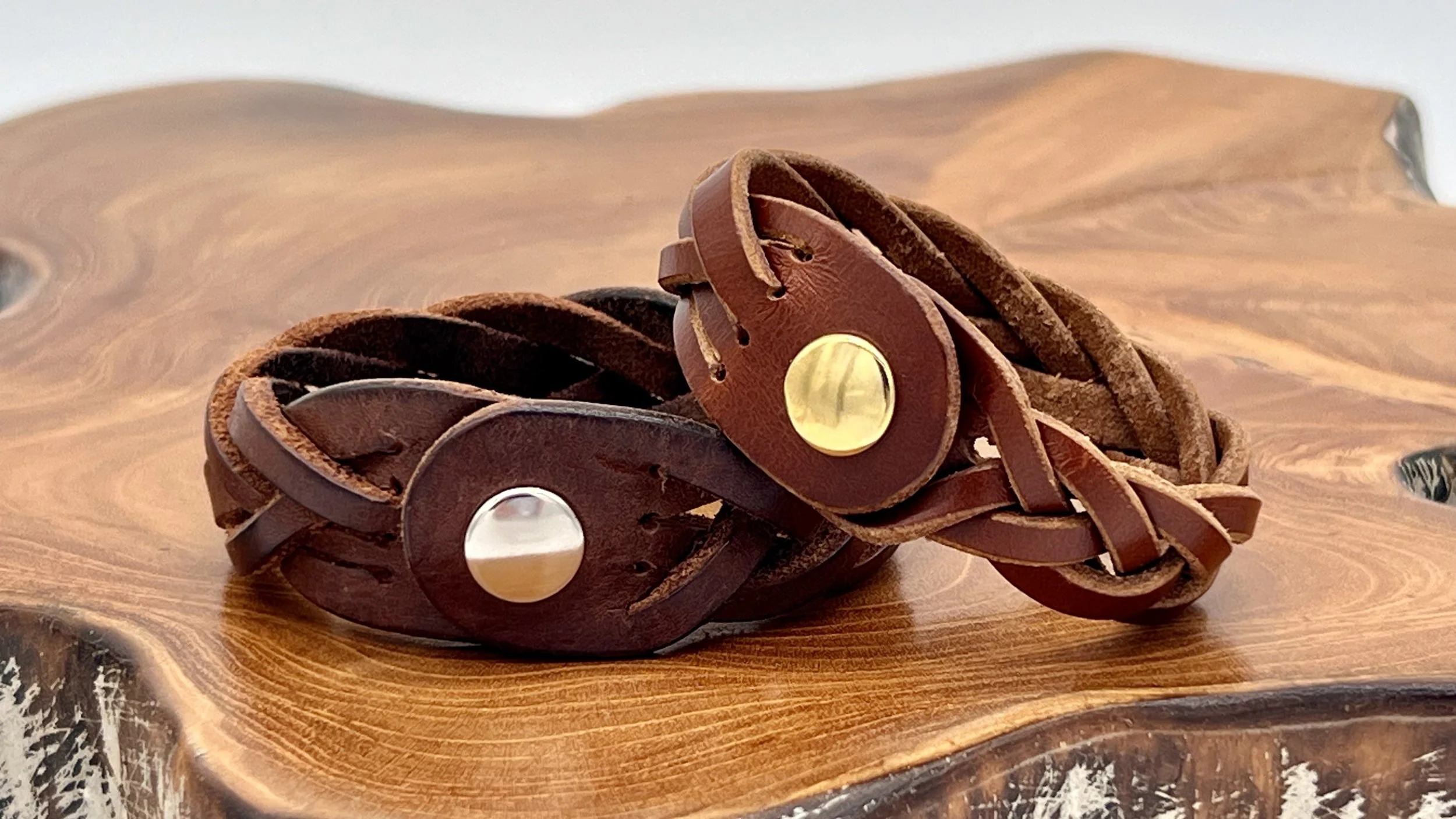 Two intertwined leather bracelets, one dark brown and the other a reddish-brown, with metallic circular embellishments, resting on a polished wooden surface.