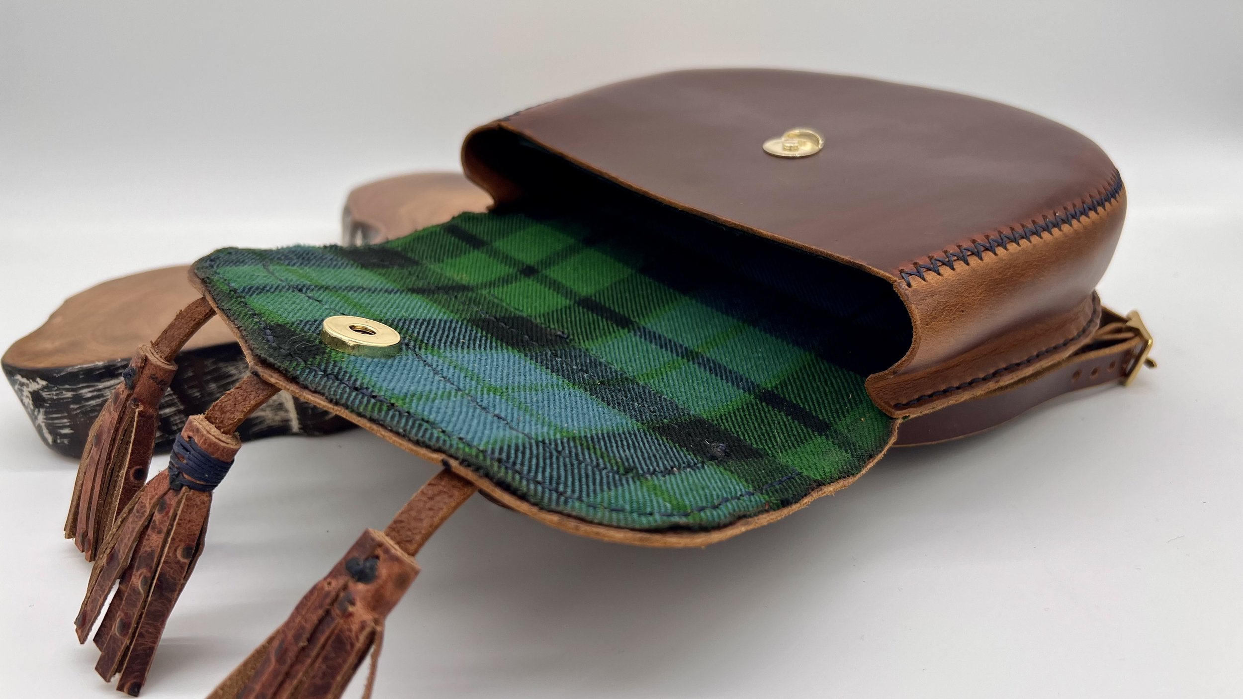 A leather handbag with a green plaid fabric interior, gold snap buttons, and leather tassels on the zipper pull, placed on a white surface.