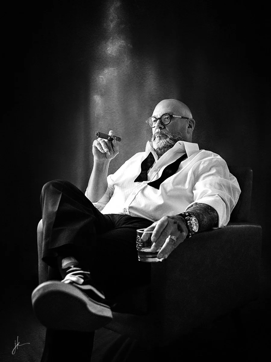 A bald man with a beard and glasses sitting in a chair, smoking a cigar, wearing a white shirt with the top buttons open, a black tie hanging loose, and dark pants. He has a watch and ring on his fingers, holding a glass of whiskey in one hand and an unlit cigar in the other.