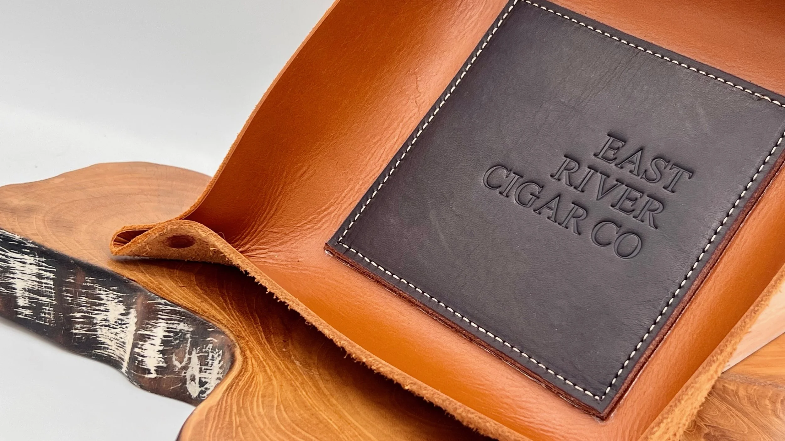 Close-up of a leather tray with a black leather patch engraved with 'EAST RIVER CIGAR CO', sitting on a wooden surface.
