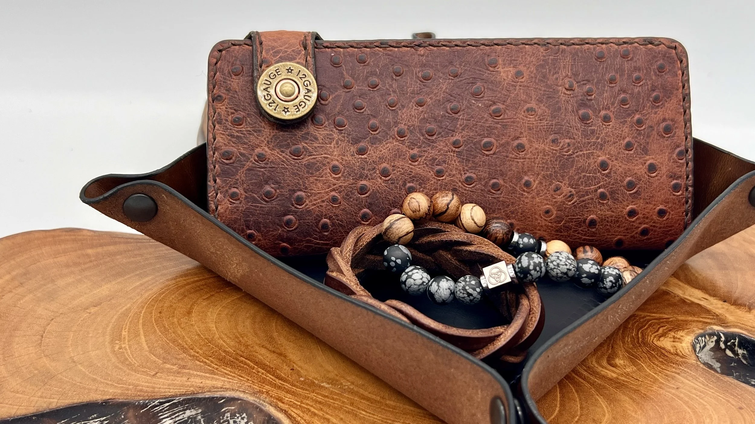 A leather tray contains a brown leather phone case with a textured surface, a beaded bracelet with black and white marble-like beads, and a wooden beaded bracelet, all placed on a wooden surface with a light background.