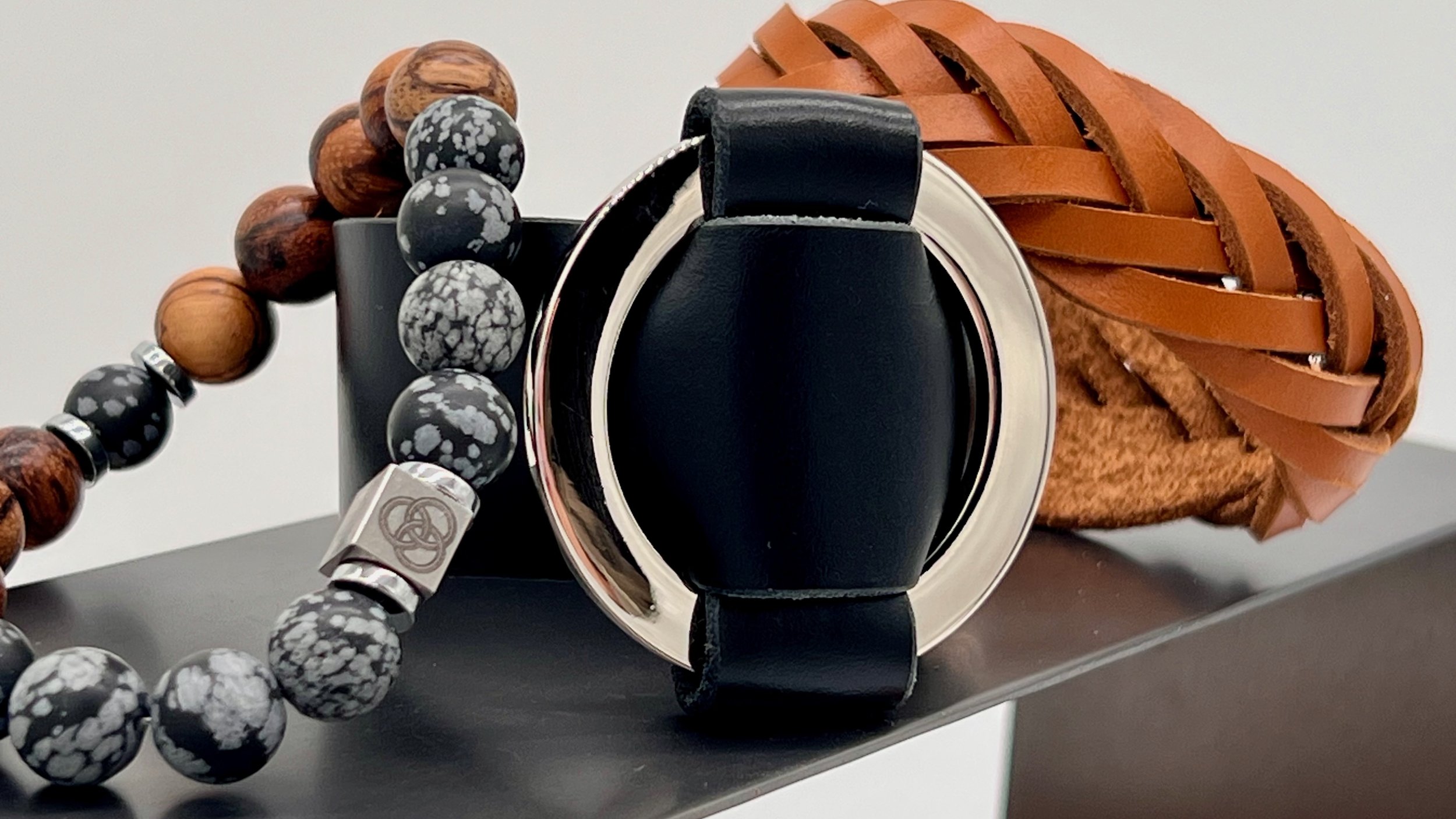 A collection of jewelry and accessories including a beaded necklace with black and gray beads, a silver ring, a black leather strap watch, and a woven brown leather clutch, all displayed on a black surface.