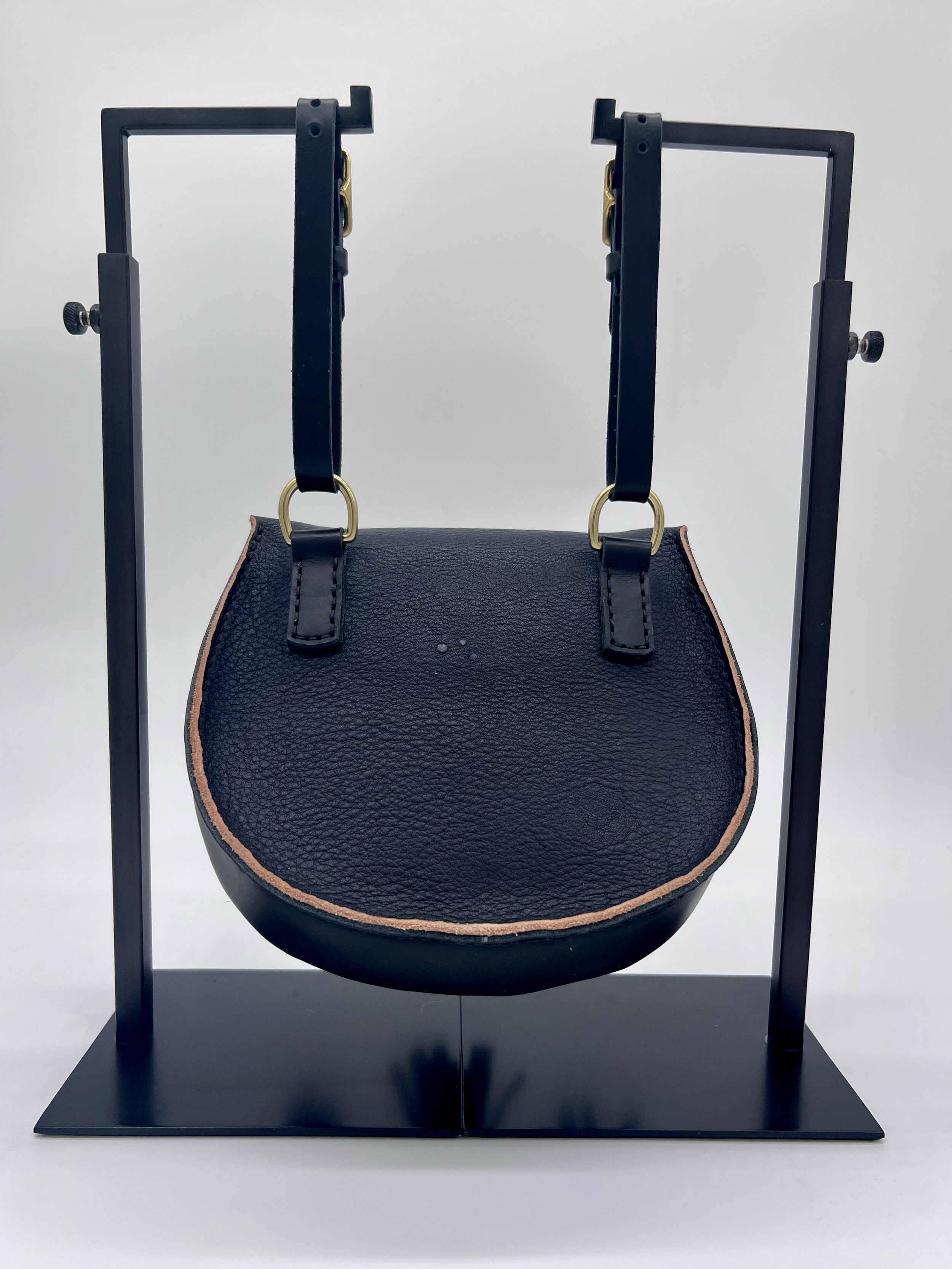 Black leather saddlebag with a tan edging, displayed on a black metal stand.