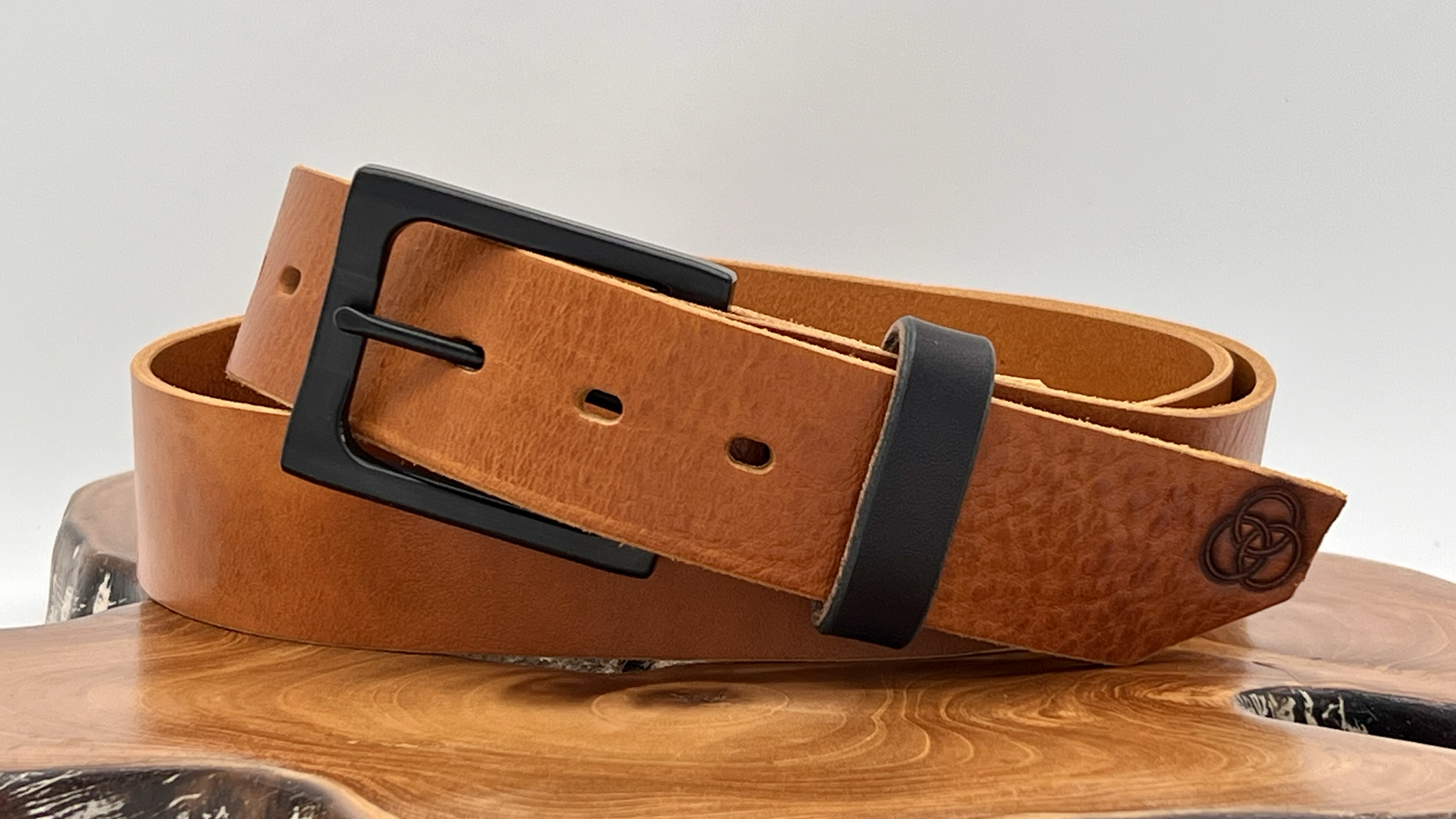 A light brown leather belt with a black buckle, placed on a wooden surface with a small knot design embossed near the end of the belt.