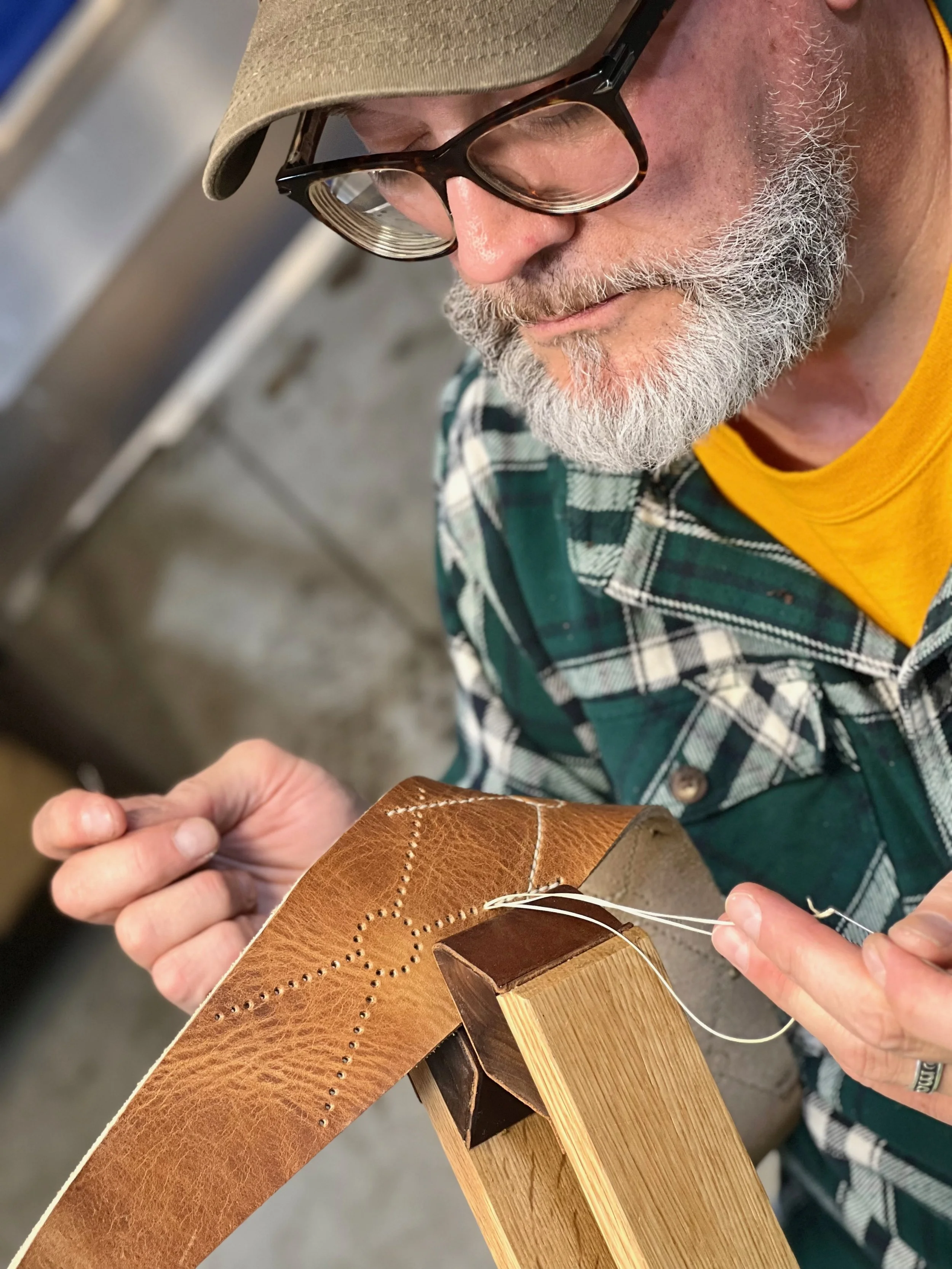 A man with glasses, a beard, and a baseball cap working on sewing leather onto a wooden piece in a workshop.