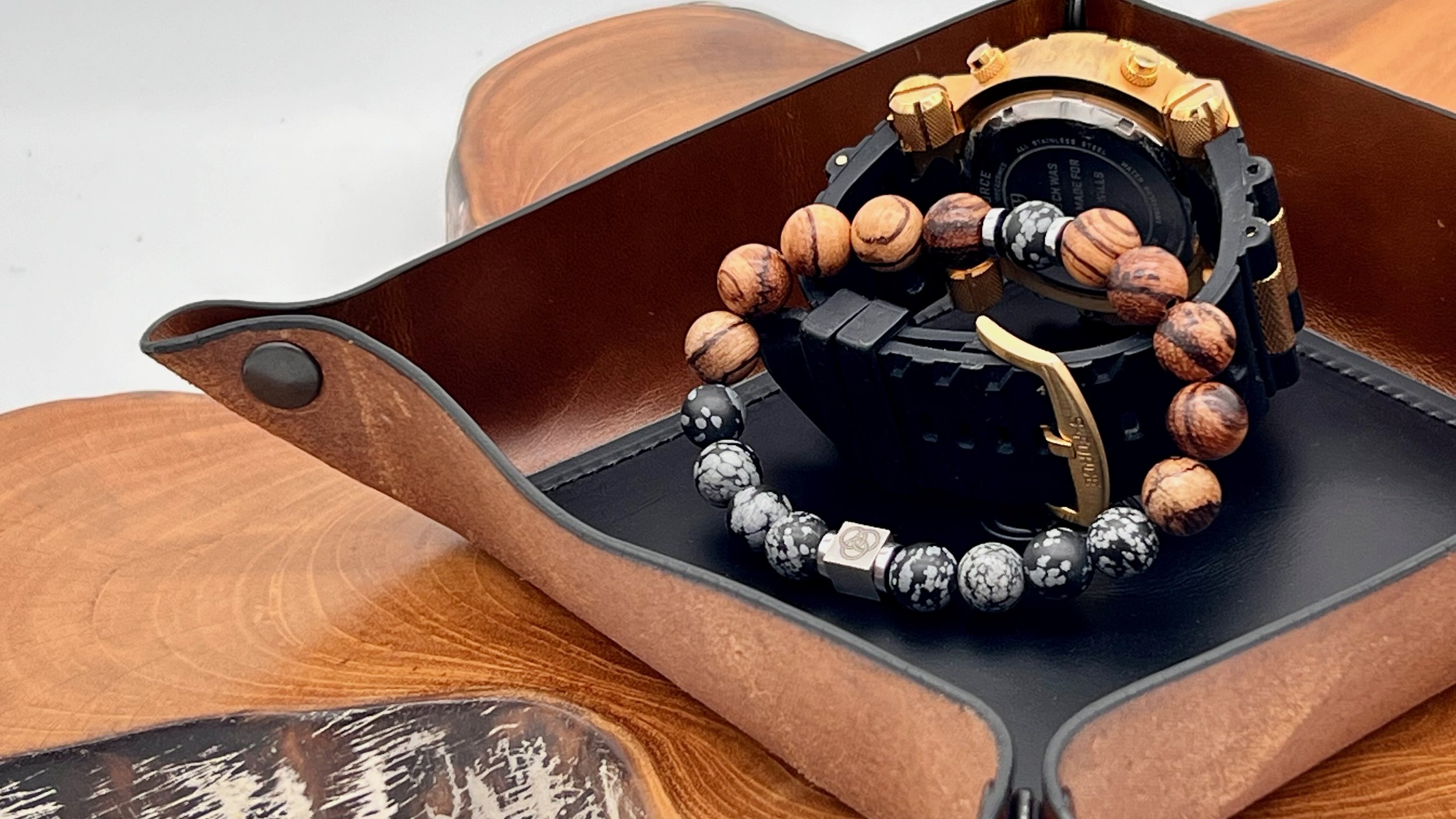 A black wristwatch with gold accents, a wooden bead bracelet, and a black and white beaded bracelet placed inside an open brown leather case on a wooden surface.